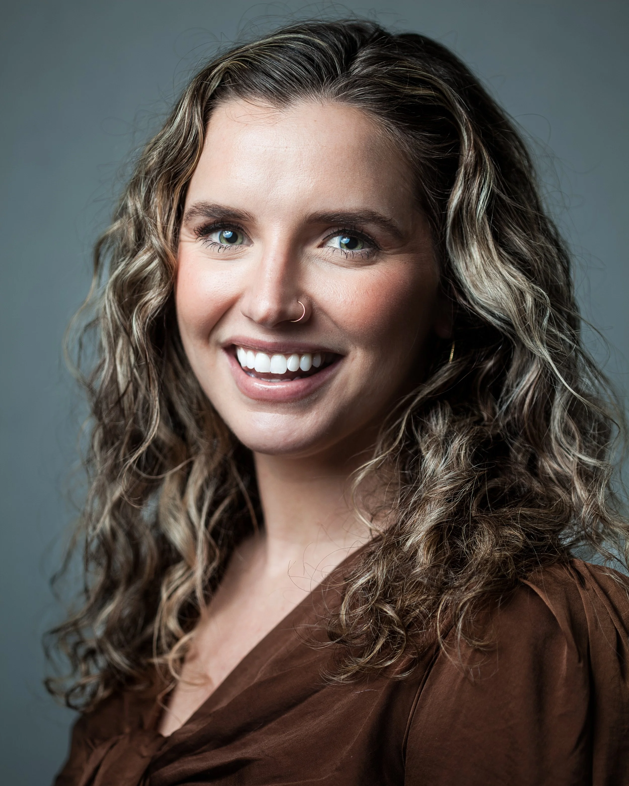 Close-up of a smiling woman with curly, shoulder-length hair, wearing a brown top and a nose ring, against a gray background.
