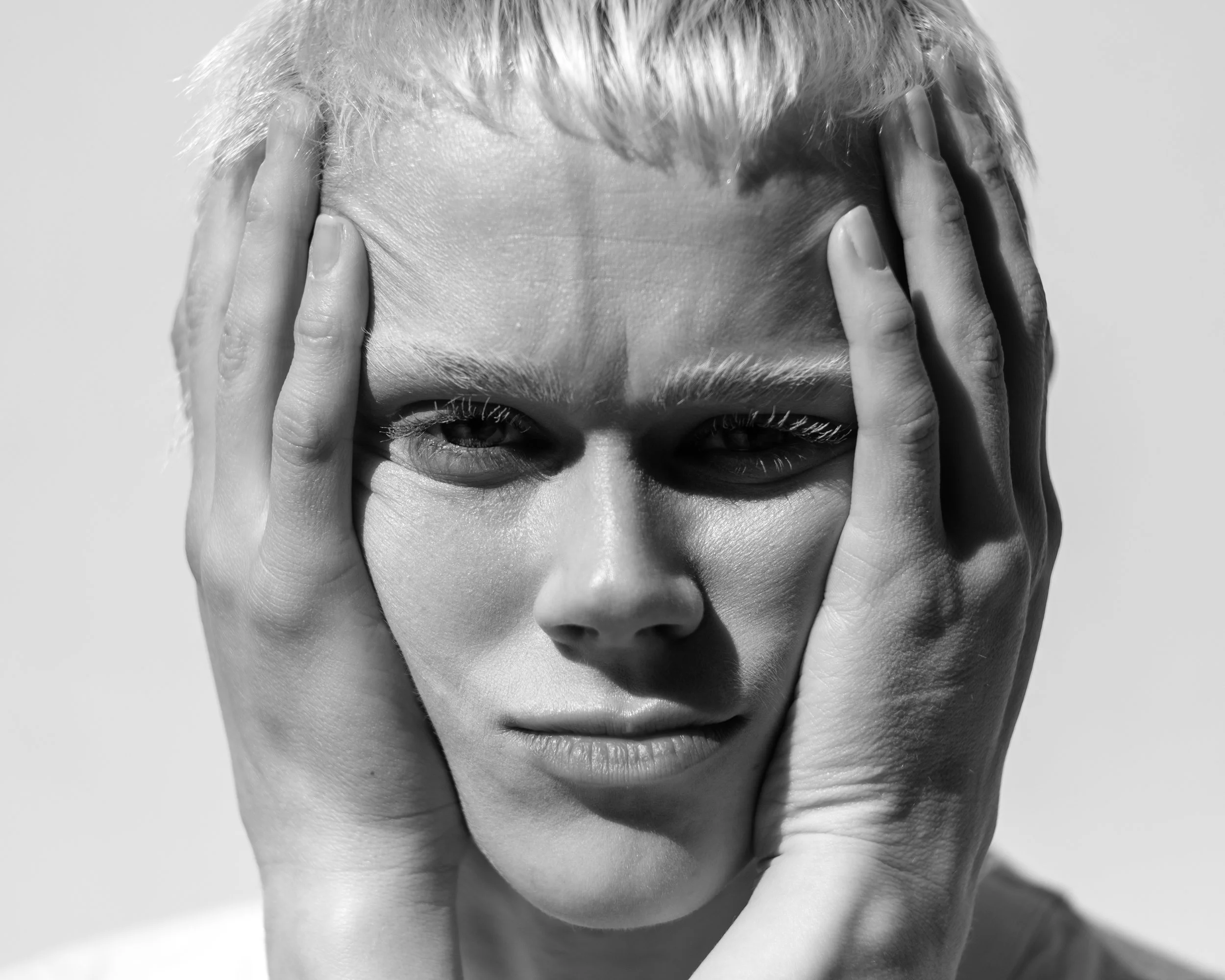 A black and white close-up of a woman with short blonde hair, holding her face with both hands, looking directly at the camera.