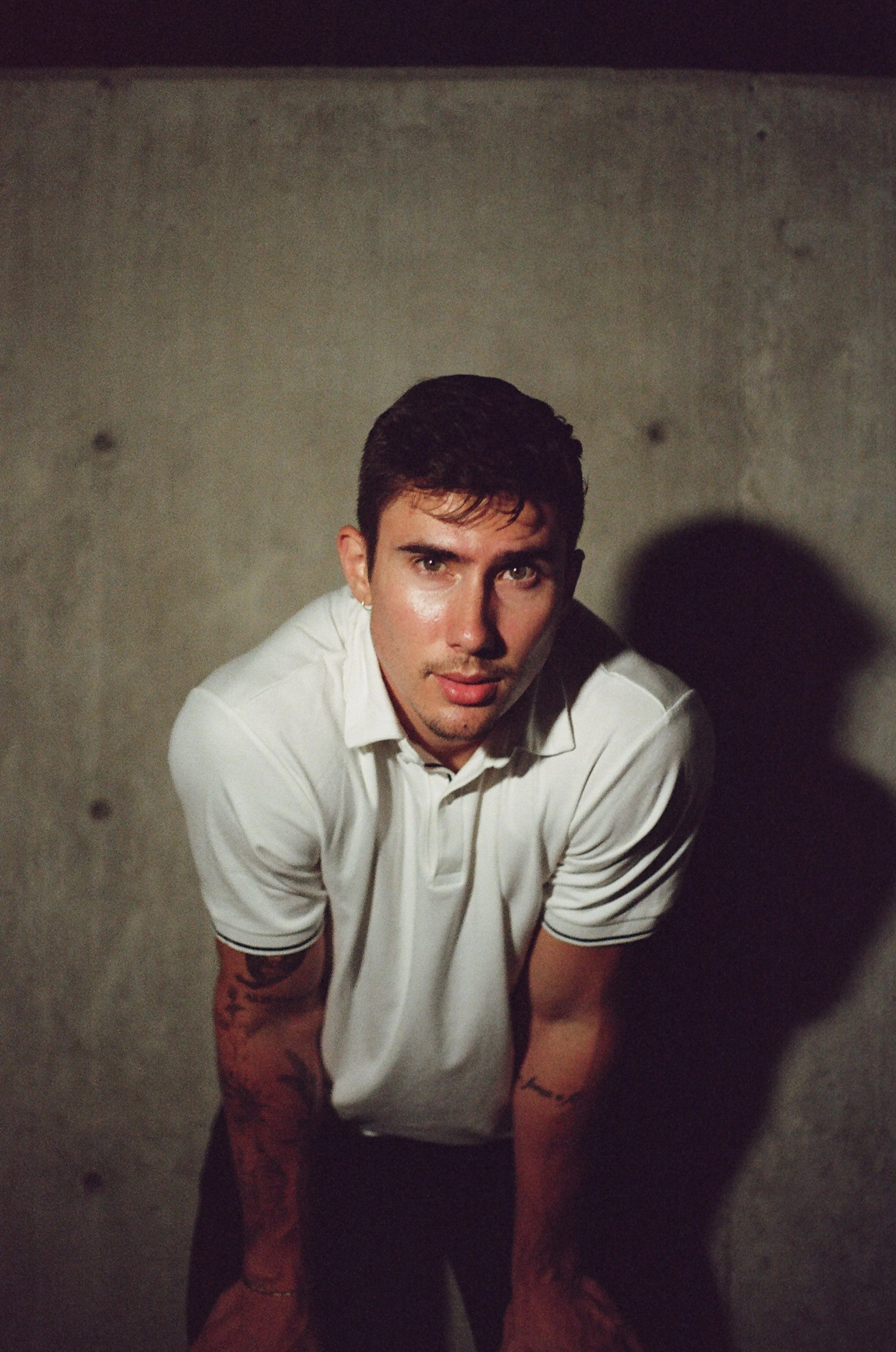 A young man with dark hair, wearing a white polo shirt, looking directly at the camera with a concrete wall in the background.