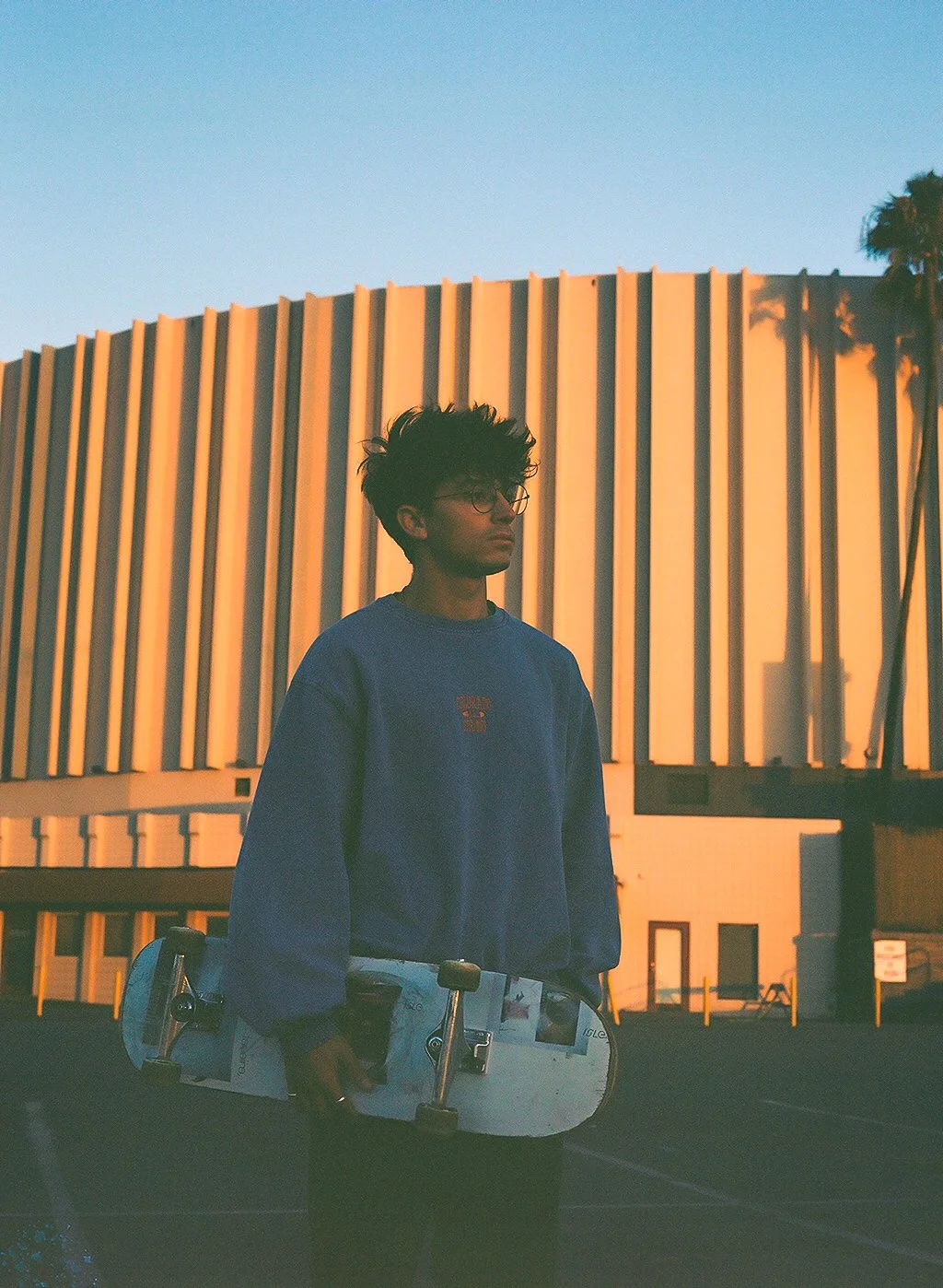 A young person with dark curly hair and glasses stands outdoors during sunset, holding a skateboard, in front of a modern, illuminated building with vertical panels.