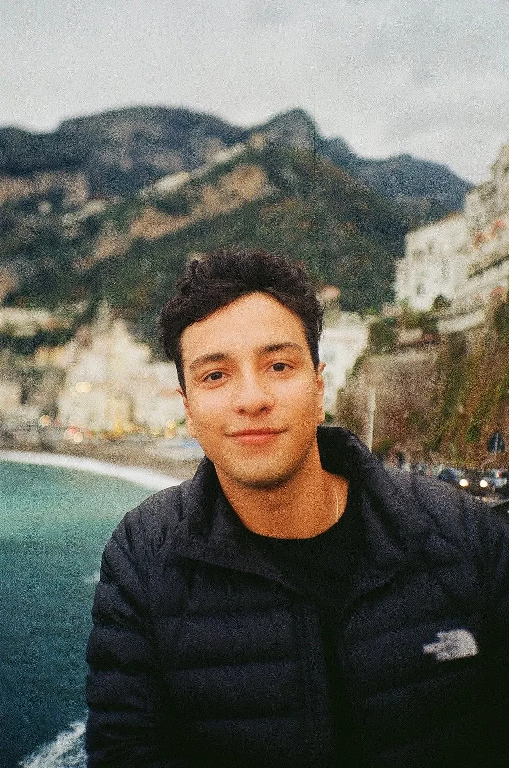 A young man with dark hair smiling in front of a scenic coastal background with cliffs and buildings, wearing a black North Face jacket.