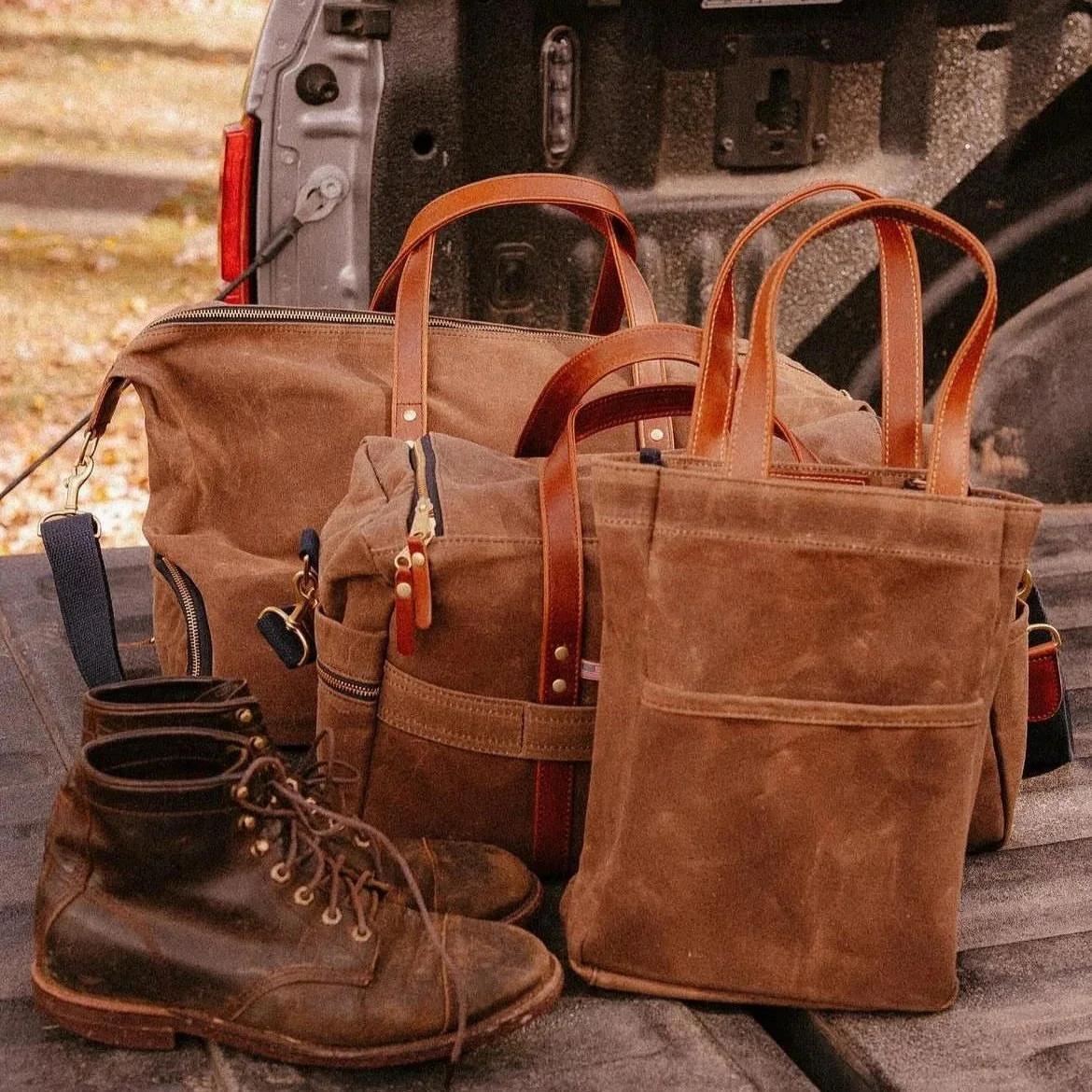 A collection of brown and tan travel bags and a pair of brown leather boots placed on a wooden surface beside a vehicle.