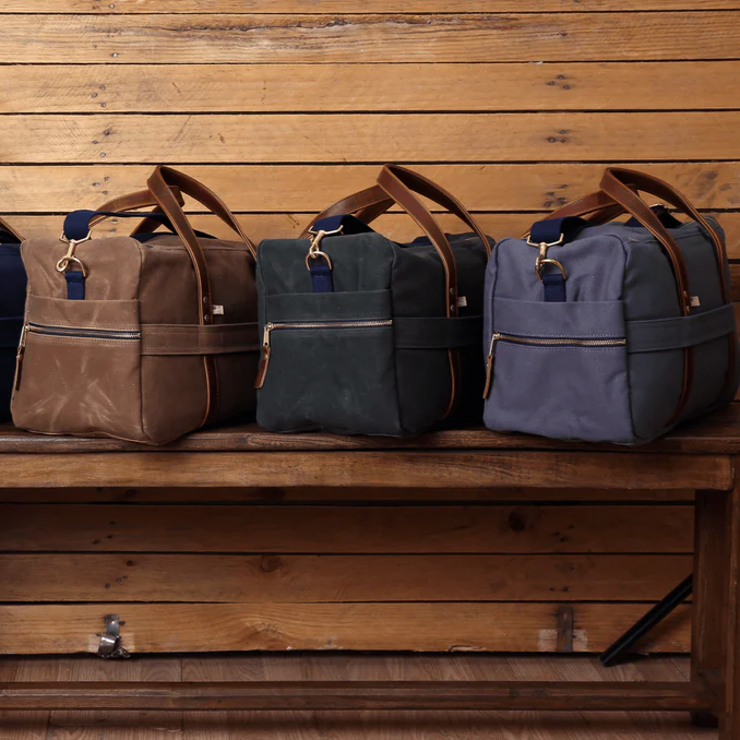 Three travel duffel bags in brown, black, and blue on a wooden bench against a wooden wall.