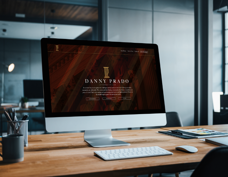 A modern office workspace with an iMac computer displaying a law firm's website called Danny Prado, with a wooden desk, keyboard, mouse, and stationery.