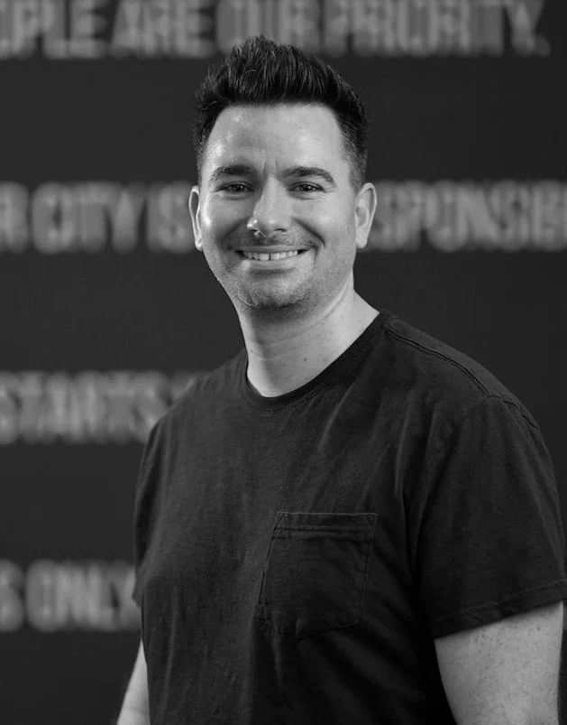 Black and white photo of a smiling man with short dark hair, wearing a plain black T-shirt, standing in front of a blurry background with text.
