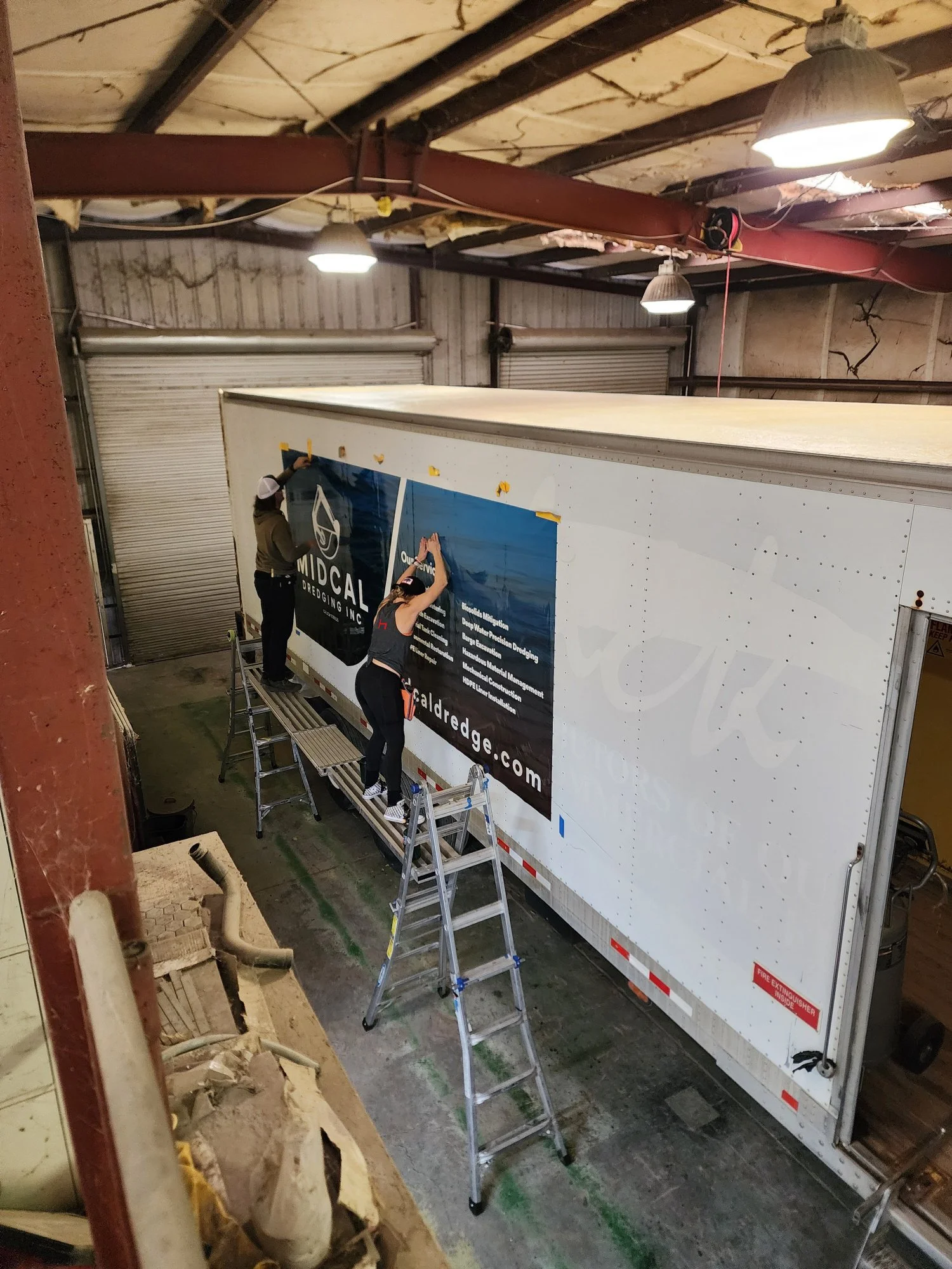 Two individuals are working on applying a banner to the side of a large white truck parked inside a warehouse. They are standing on ladders, attaching the banner that displays a logo, website, and various texts.