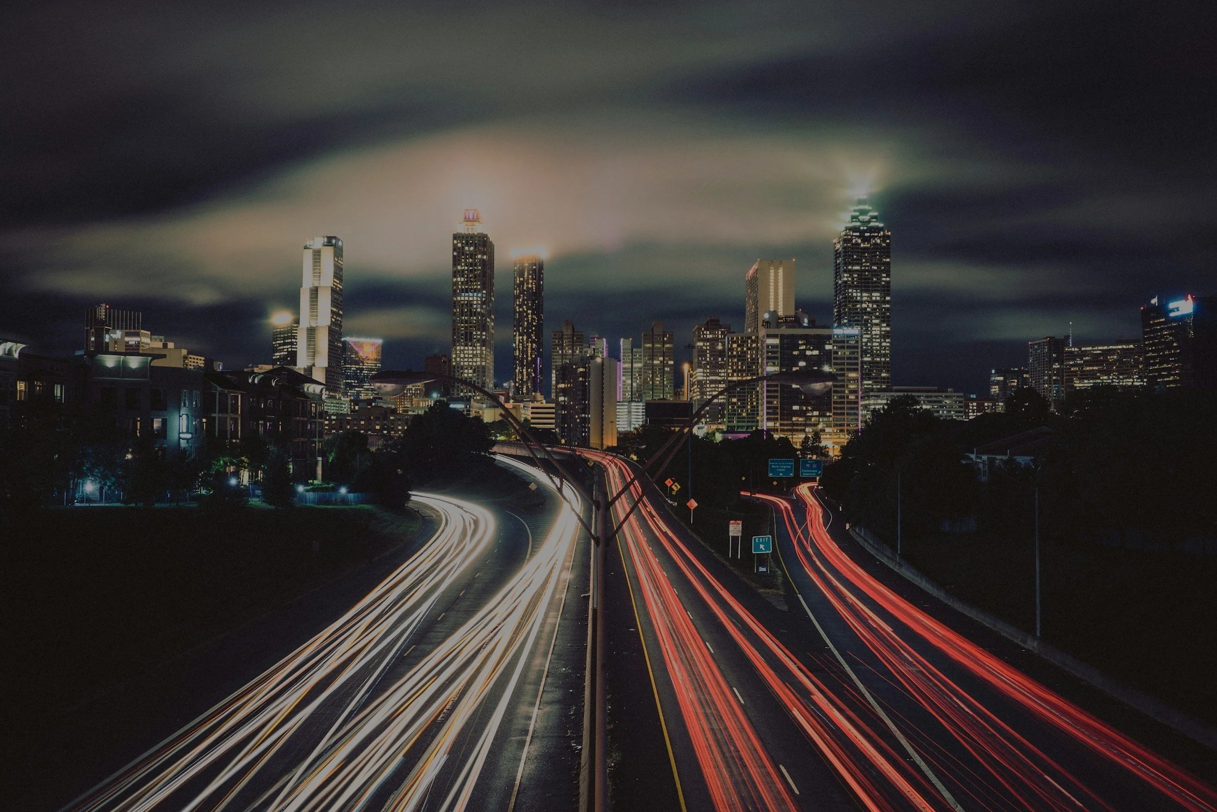 Nighttime Atlanta skyline featuring illuminated skyscrapers under a cloudy sky with visible city traffic light trails on roads.