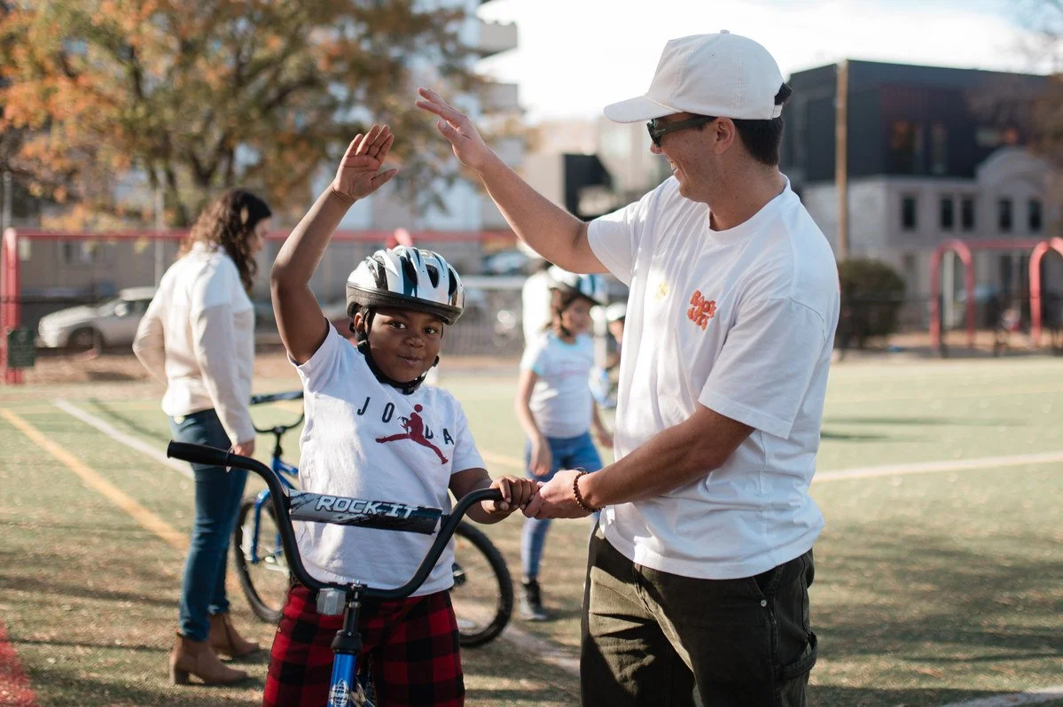 Child giving a high five to a volunteer while riding a bicycle at a community event