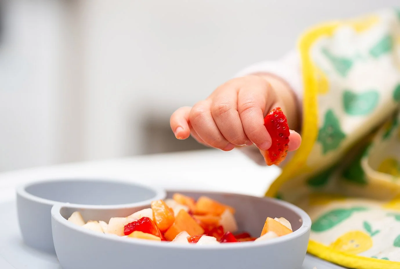 Close-up of toddler holding a piece of strawberry above a bowl of chopped fruit during snack time