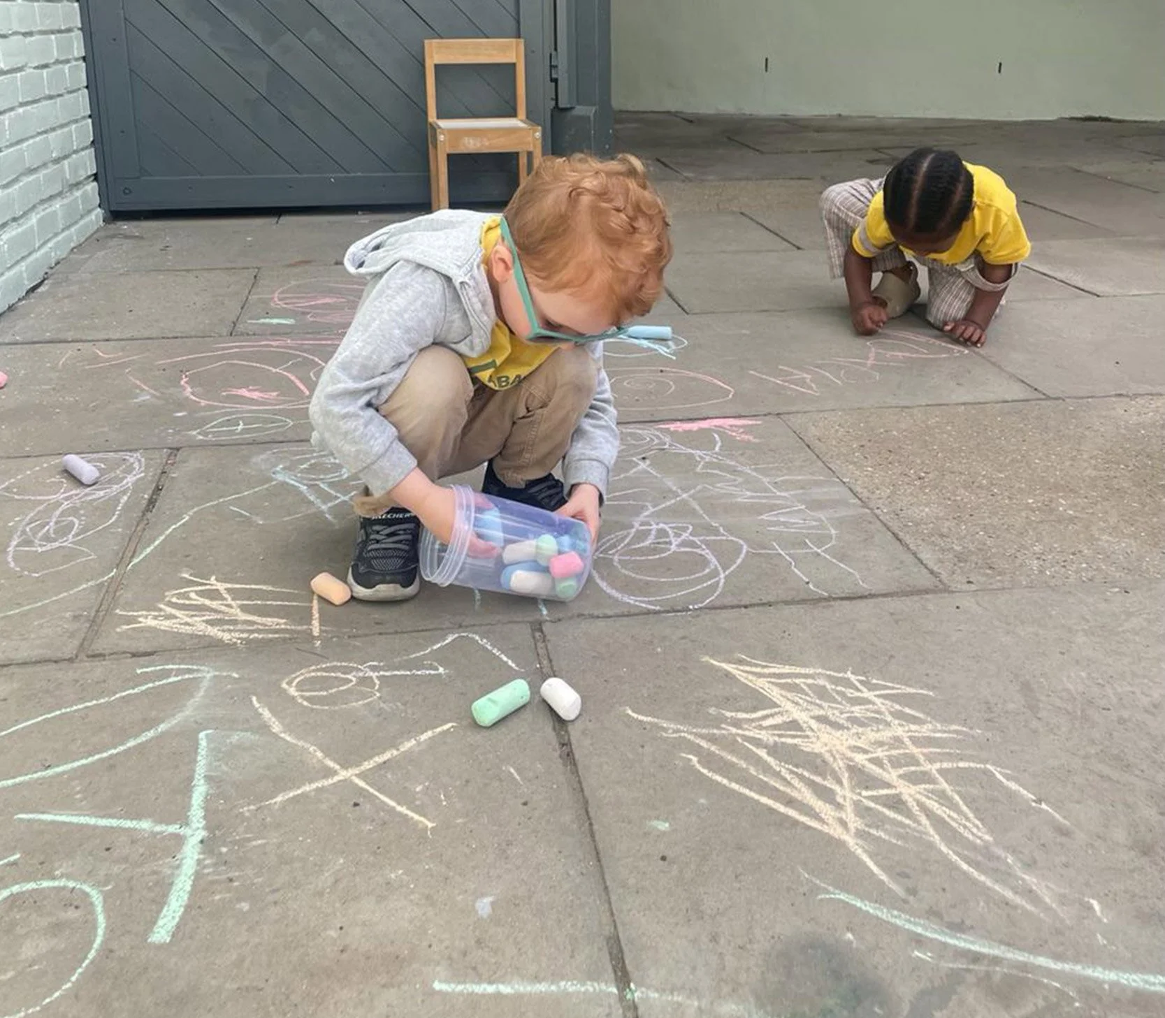 Children developing fine motor skills through outdoor mark-making with large sidewalk chalks.