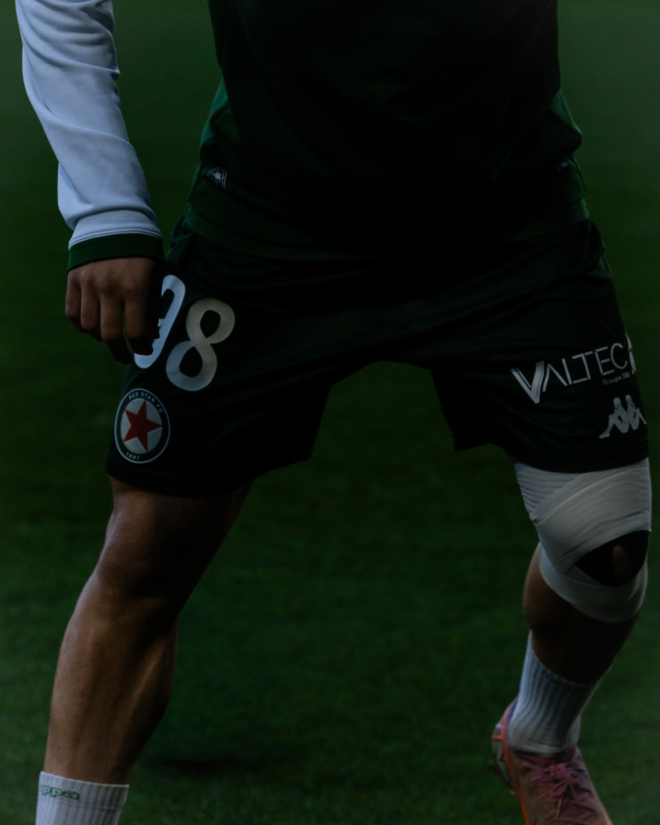 Un joueur de football en position sur le terrain, portant un maillot noir avec des logos et des numéros blancs, une jambe blessée avec un bandage blanc, et des chaussures de sport rose et orange.