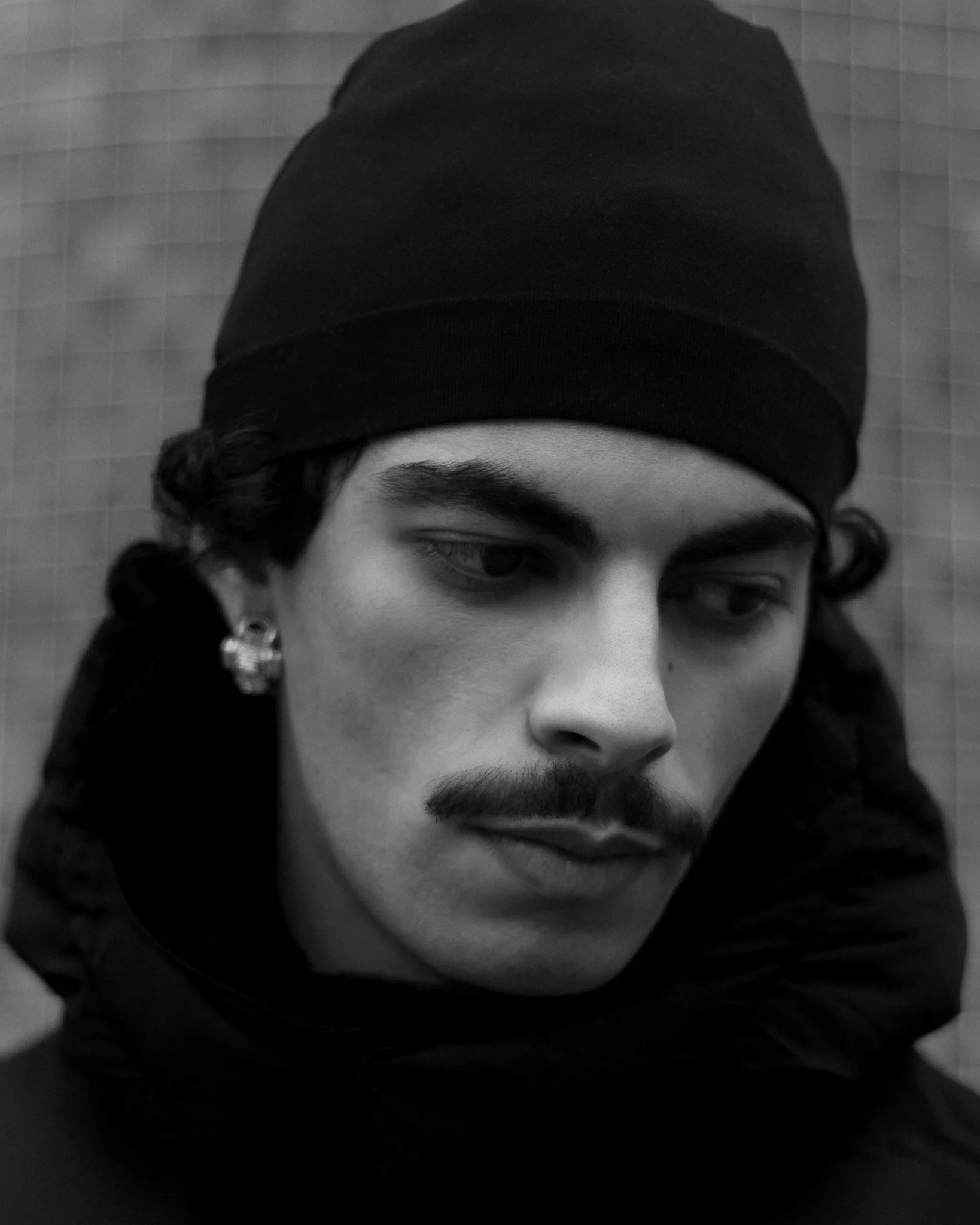 Portrait en noir et blanc d'un jeune homme avec un bandeau noir sur la tête, des boucles d'oreilles, un cache-cou noir et une moustache, regardant vers le bas.