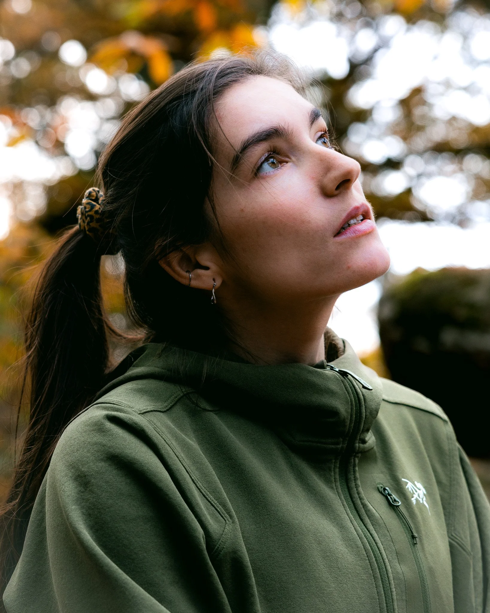 Jeune femme regardant vers le haut, portant une veste verte, fond d'arbres aux couleurs automnales.