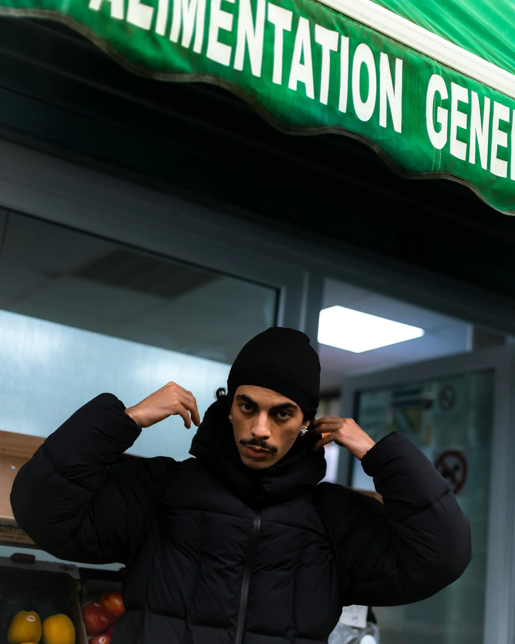 Un jeune homme en tenue noire ajuste ses écouteurs à l'intérieur d'un magasin, près d'un panneau vert avec des lettres en blanc, évoquant un service d'alimentation ou de nutrition.