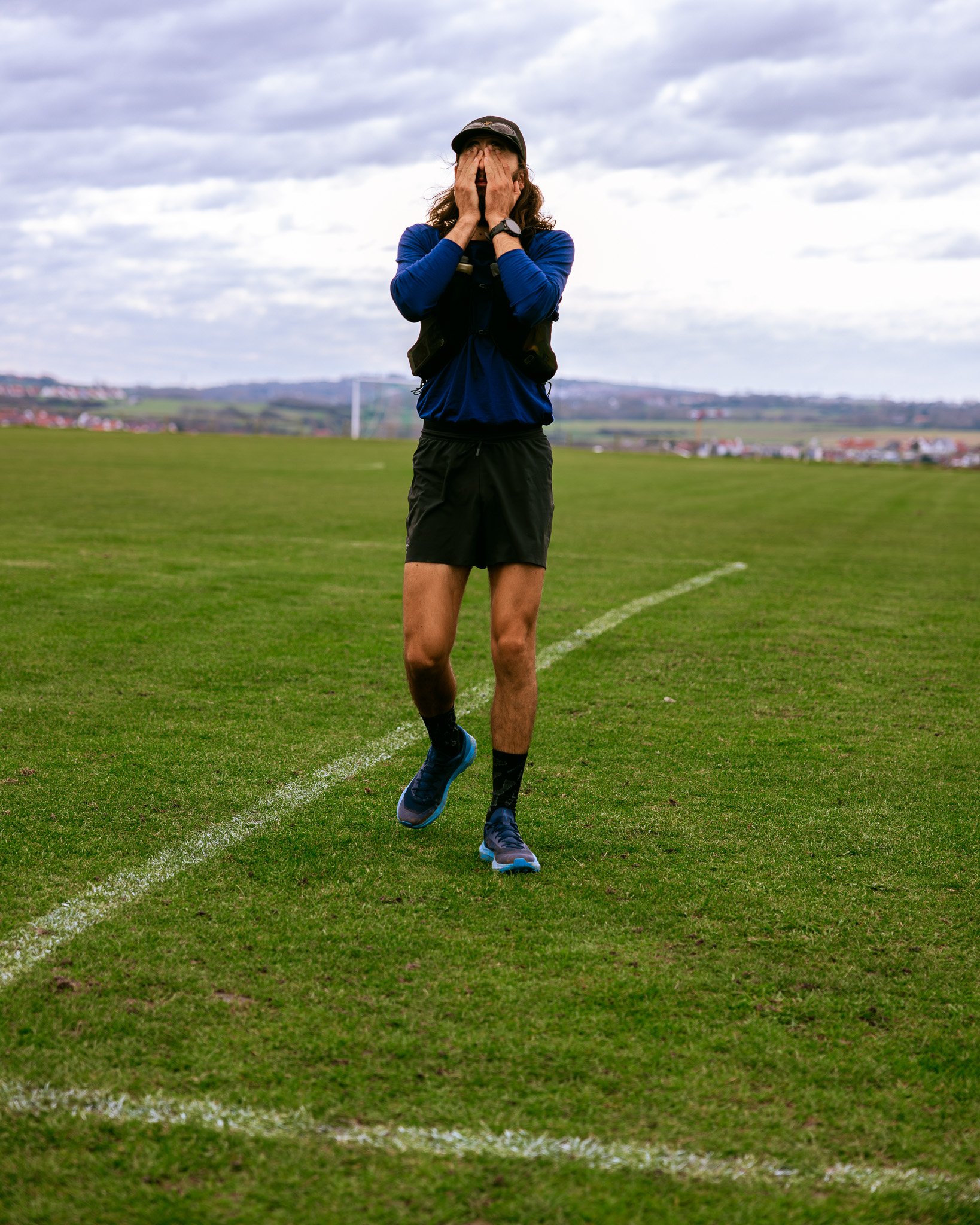 Un homme en tenue de course, portant une casquette et des chaussures de sport, est en train de courir sur un terrain de sport en plein air, sa main couvrant son visage.