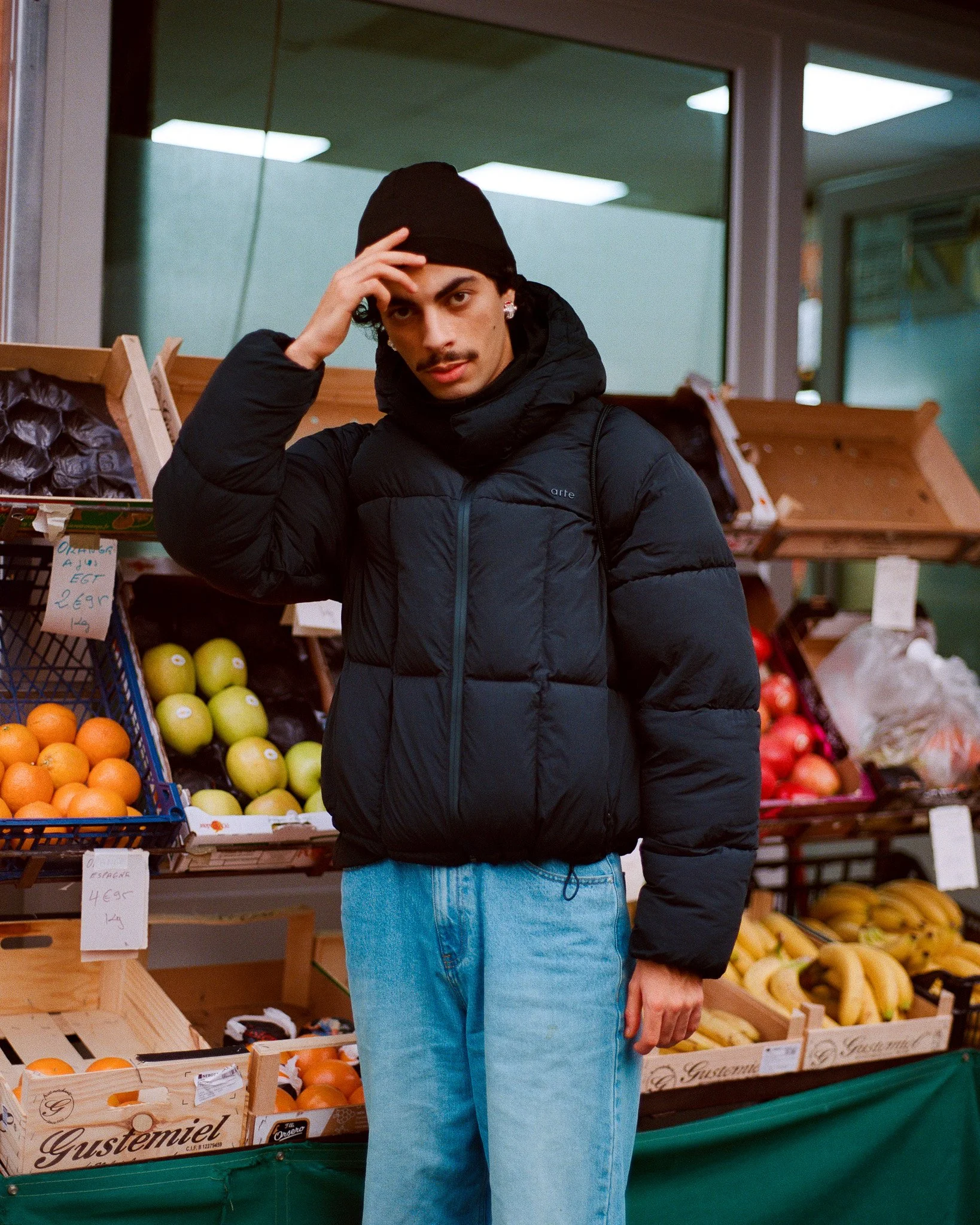 Jeune homme posant dans un marché de fruits, portant une doudoune noire, un bonnet noir, et des jeans bleus, avec des étals de fruits en arrière-plan.