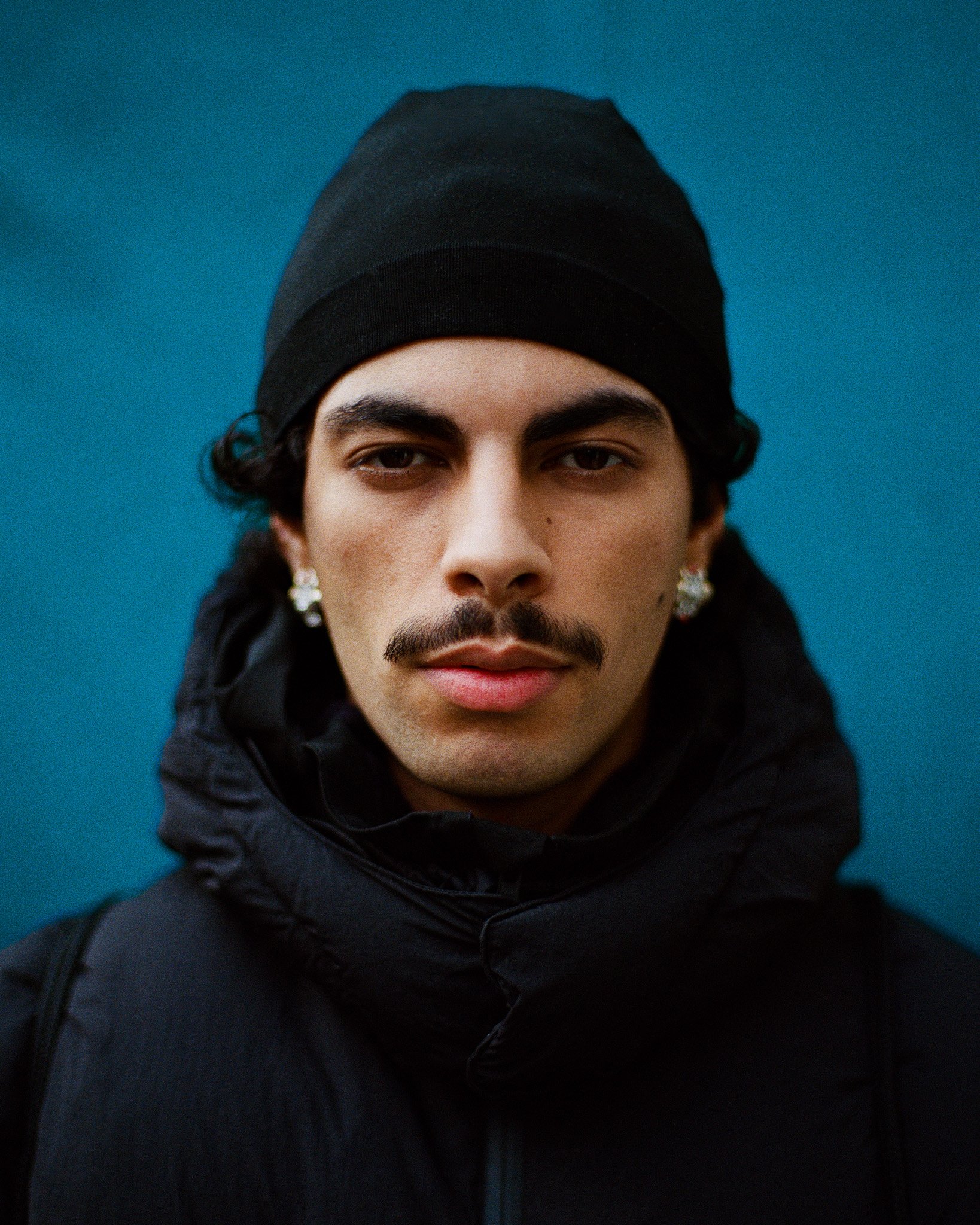 Portrait d'un jeune homme portant un bonnet noir, une veste noire et des boucles d'oreilles avec un fond bleu.
