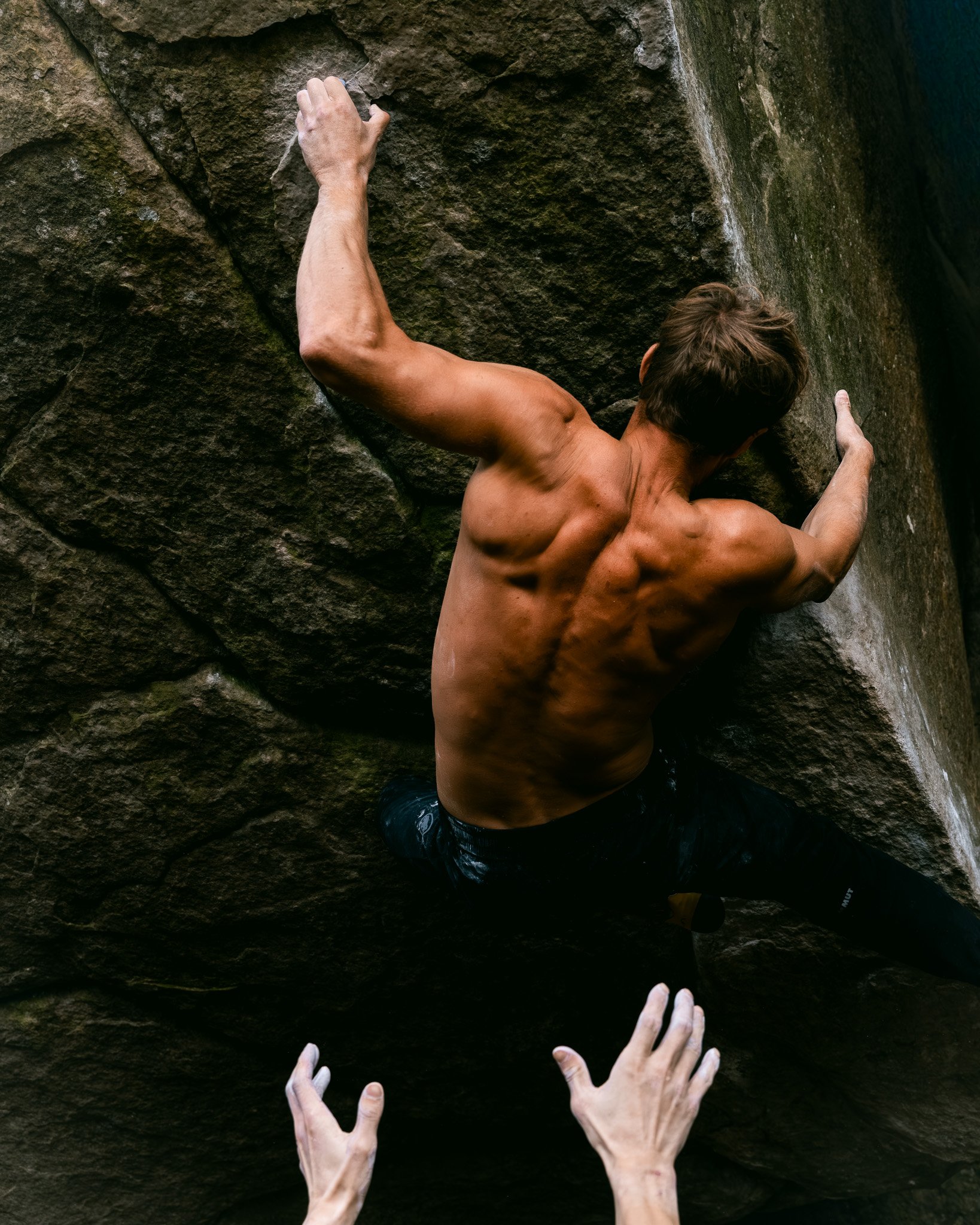 Une photographie d'un grimpeur dans la forêt de Fontainebleau. Il travaille ici un bloc de cotation élevée, la veille du conte. Homme en pleine escalade de rochers, vu de dos, avec mains visibles en contre-plongée.