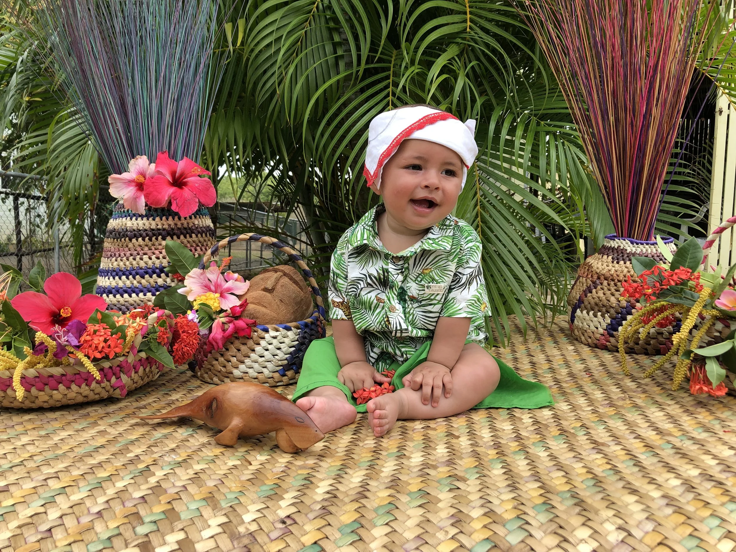 An image of a baby boy at his first haircut ceremony on Boigu Island. He is wearing his totem colour green, with beautiful handmade baskets, tropical flowers and carvings decorating his photoshoot.