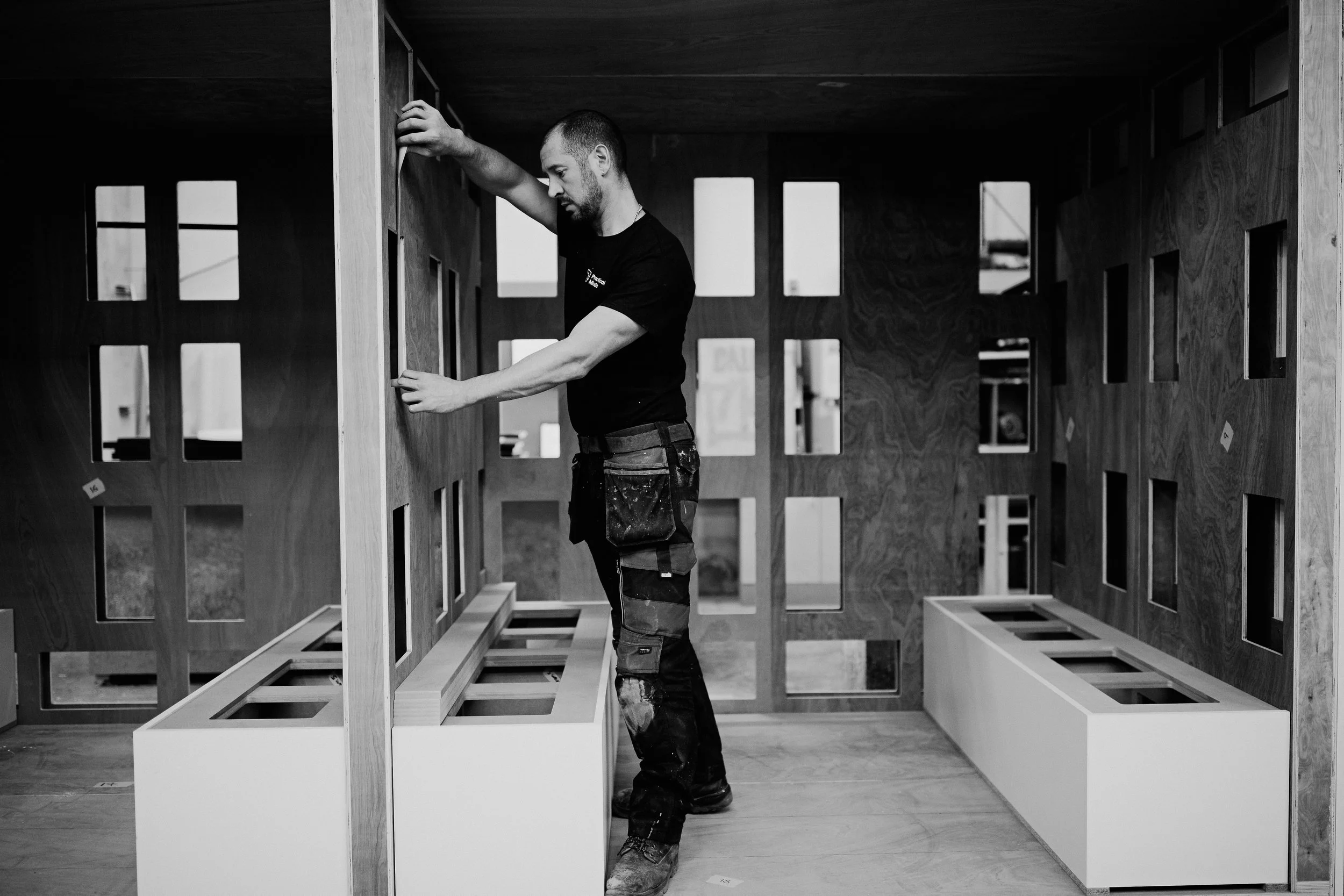 A man working inside a partially constructed wooden structure, adjusting a wooden panel with cut-out sections. He is wearing a tool belt and appears to be engaged in carpentry or construction work.