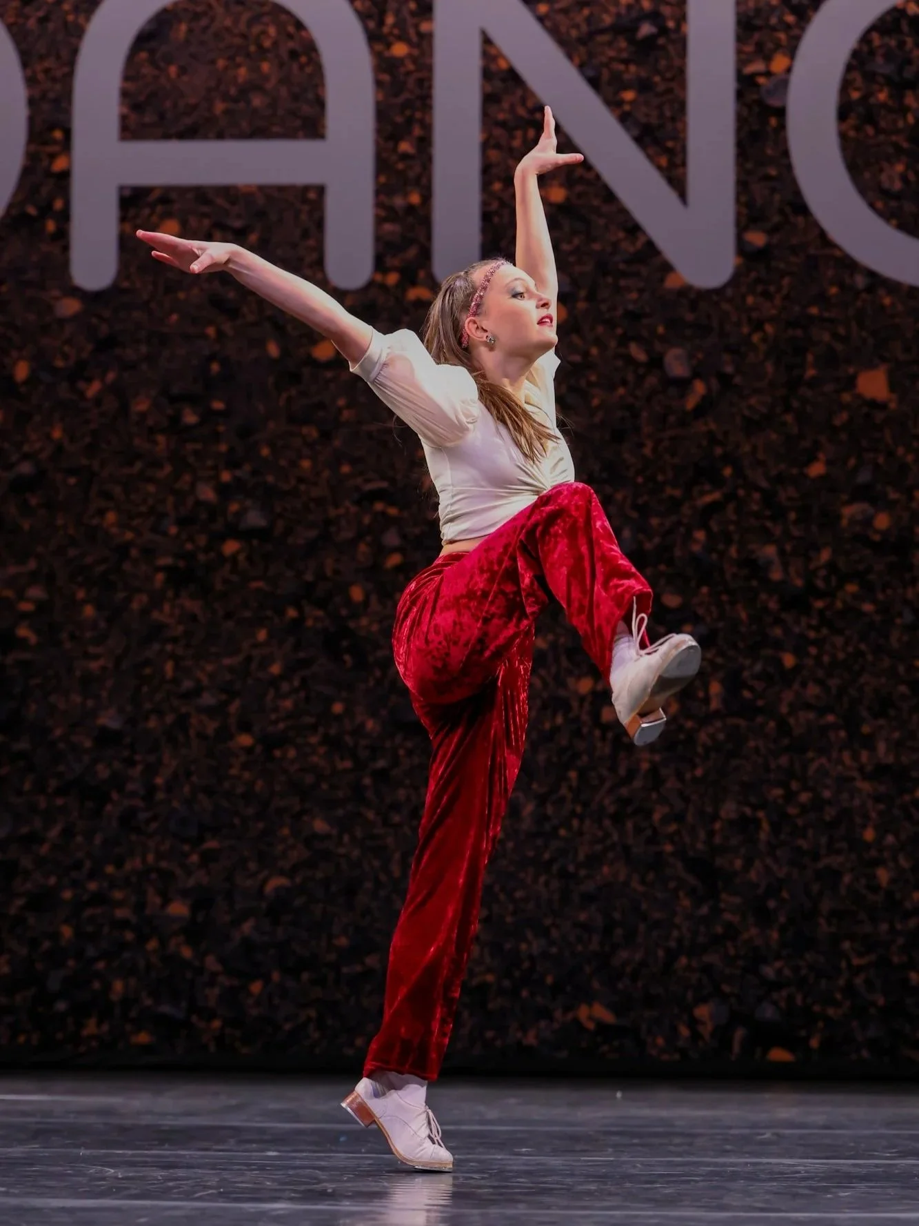 A female dancer performing on stage with a dark, textured backdrop and large letters in the background. She is balancing on one leg with her arms extended, wearing a beige top, red velvet pants, and white dance tap shoes. i am DANCE Competition