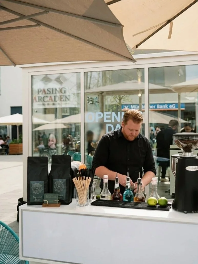 Barista bereitet an einer mobilen Kaffeebar vor einem Café verschiedene Getränke mit Sirups, Limetten und Kaffeemaschine zu.