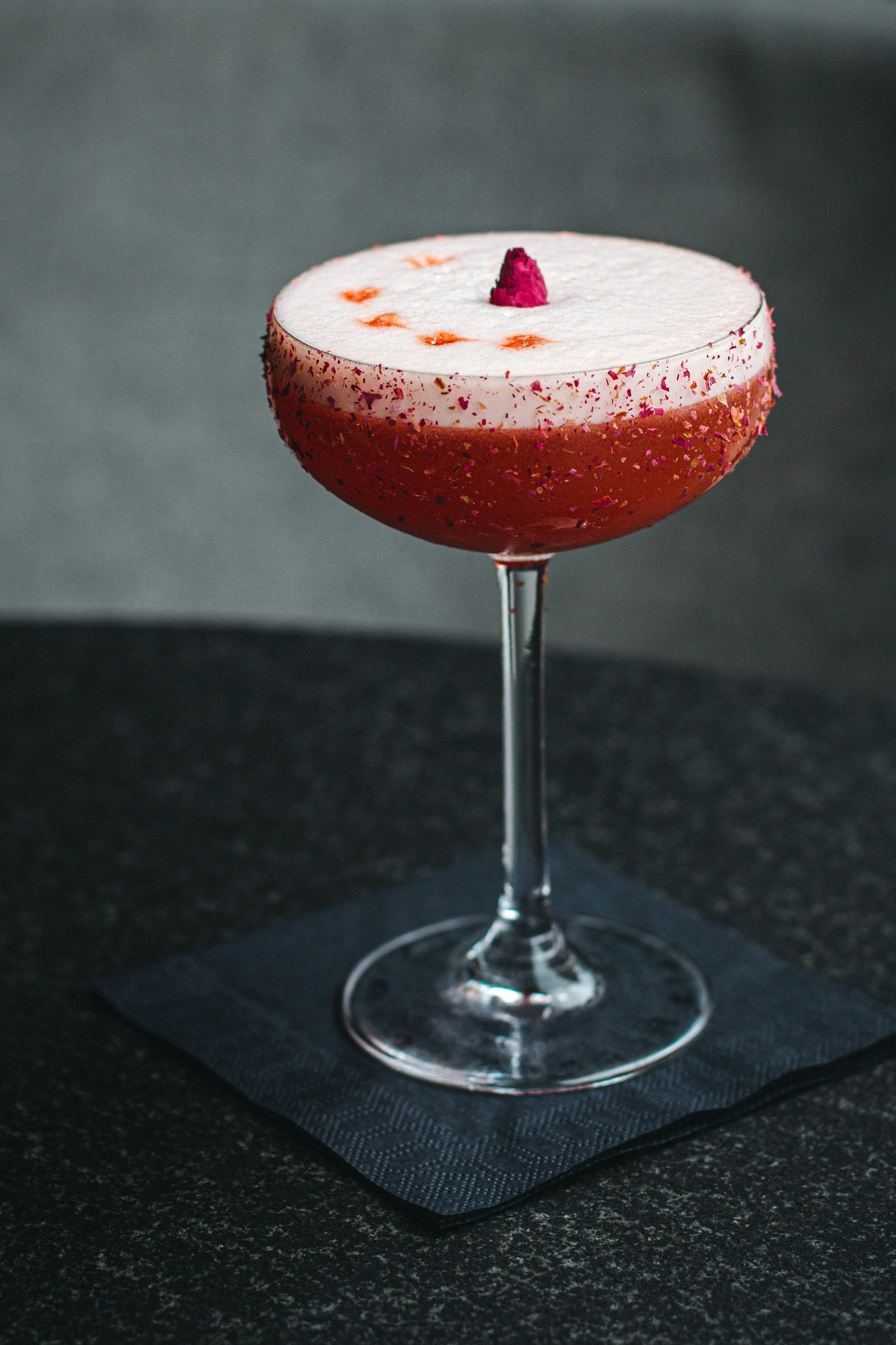 Ein Cocktail in einem gläsernen Schüssel, mit weißer Oberfläche, roten Sprenkeln, einer rosa Dekoration in der Mitte und einem Orange-Tipp, auf einem schwarzen Untersetzer auf einer dunklen Oberfläche.