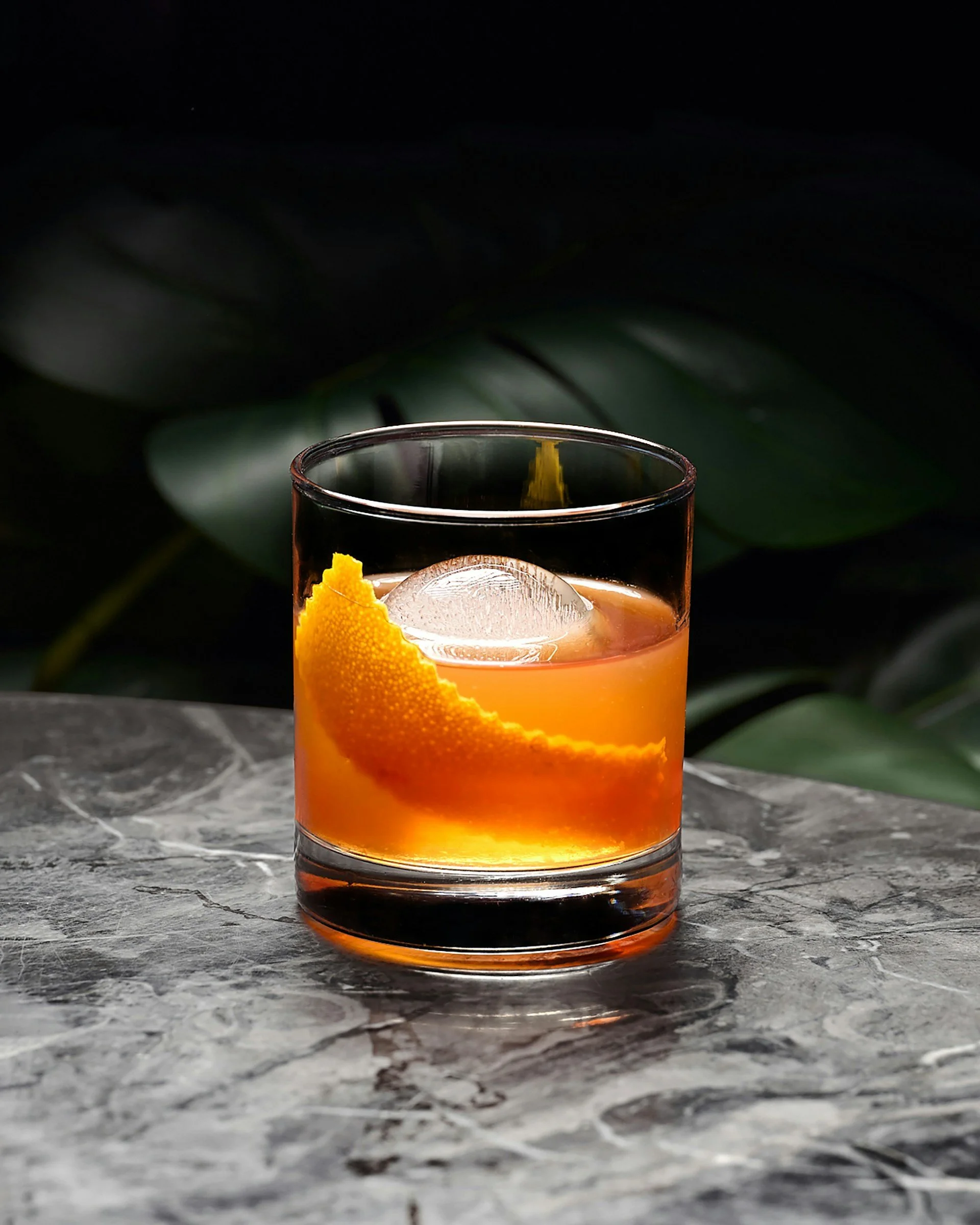 Ein Glas mit orangefarbigem Getränk, garniert mit einer Orangenschale, auf einem grauen, marmorierten Tisch vor einem dunkelgrünen Blatt im Hintergrund.