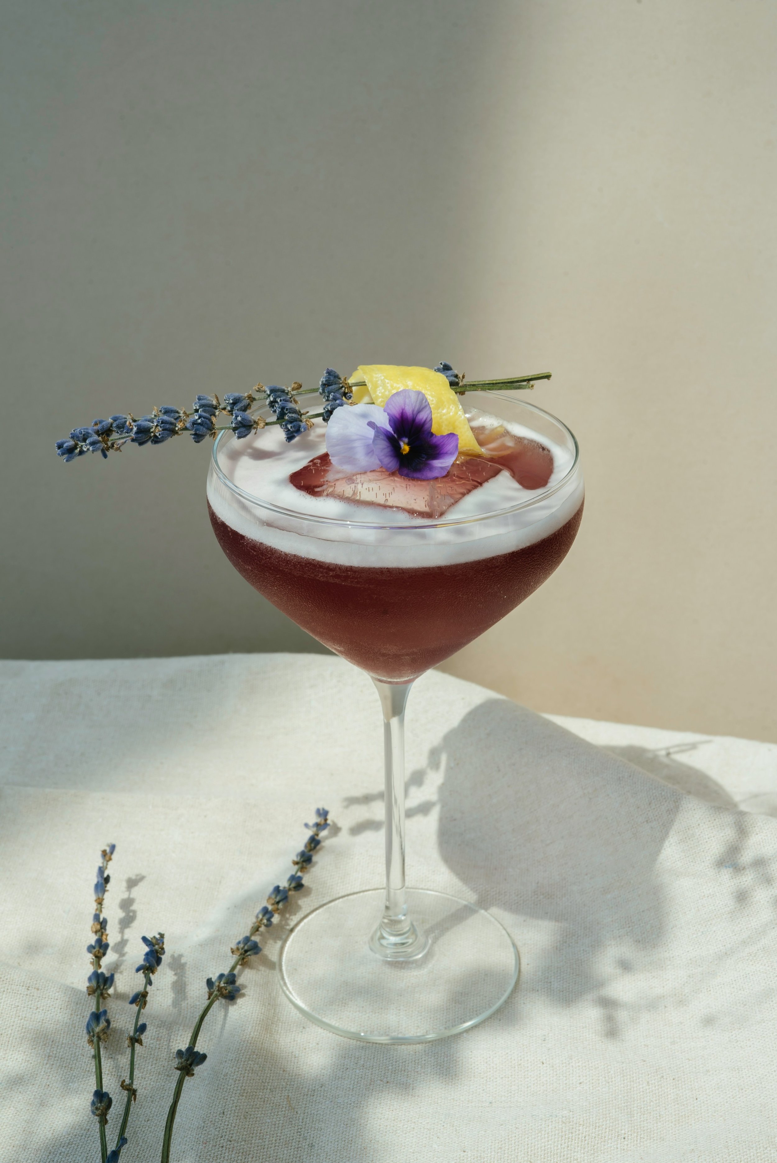 Ein Cocktail in einem Martini-Glas, dekoriert mit Lavendel, einer Zitronenscheibe und einer essbaren Blume, auf einem weißen Tuch mit Lavendelzweigen.