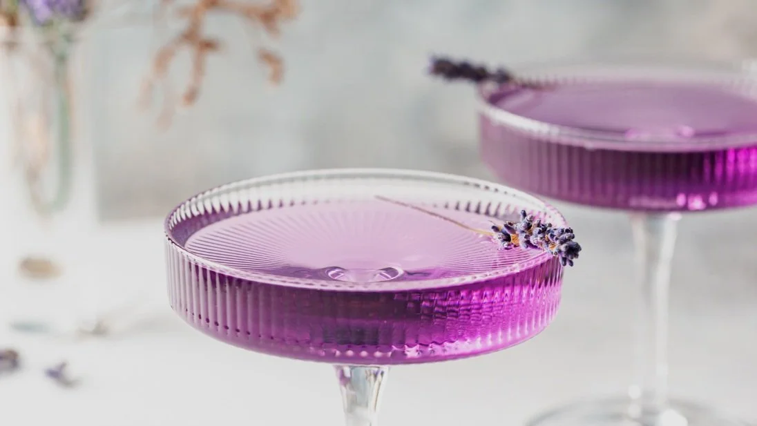 Zwei rosa-violette Cocktail-Gläser mit Lavendeldekoration auf einem weißen Tisch.