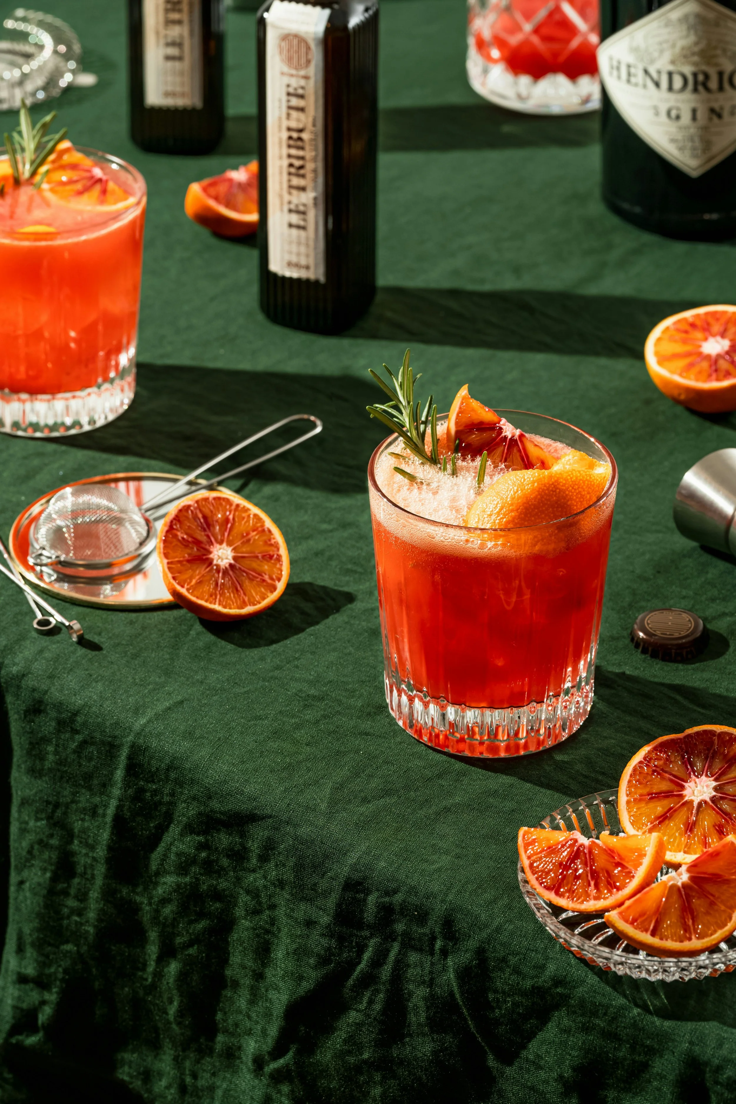 Ein Glas mit einem rosafarbenen Cocktail, garniert mit einer Rosmarin- und Blutorangen-Scheibe. Auf dem Tisch liegen halbe Blutorangen und eine Schale mit Orangenscheiben. Im Hintergrund sind Flaschen, darunter Hendrick's Gin und andere Spirituosen.