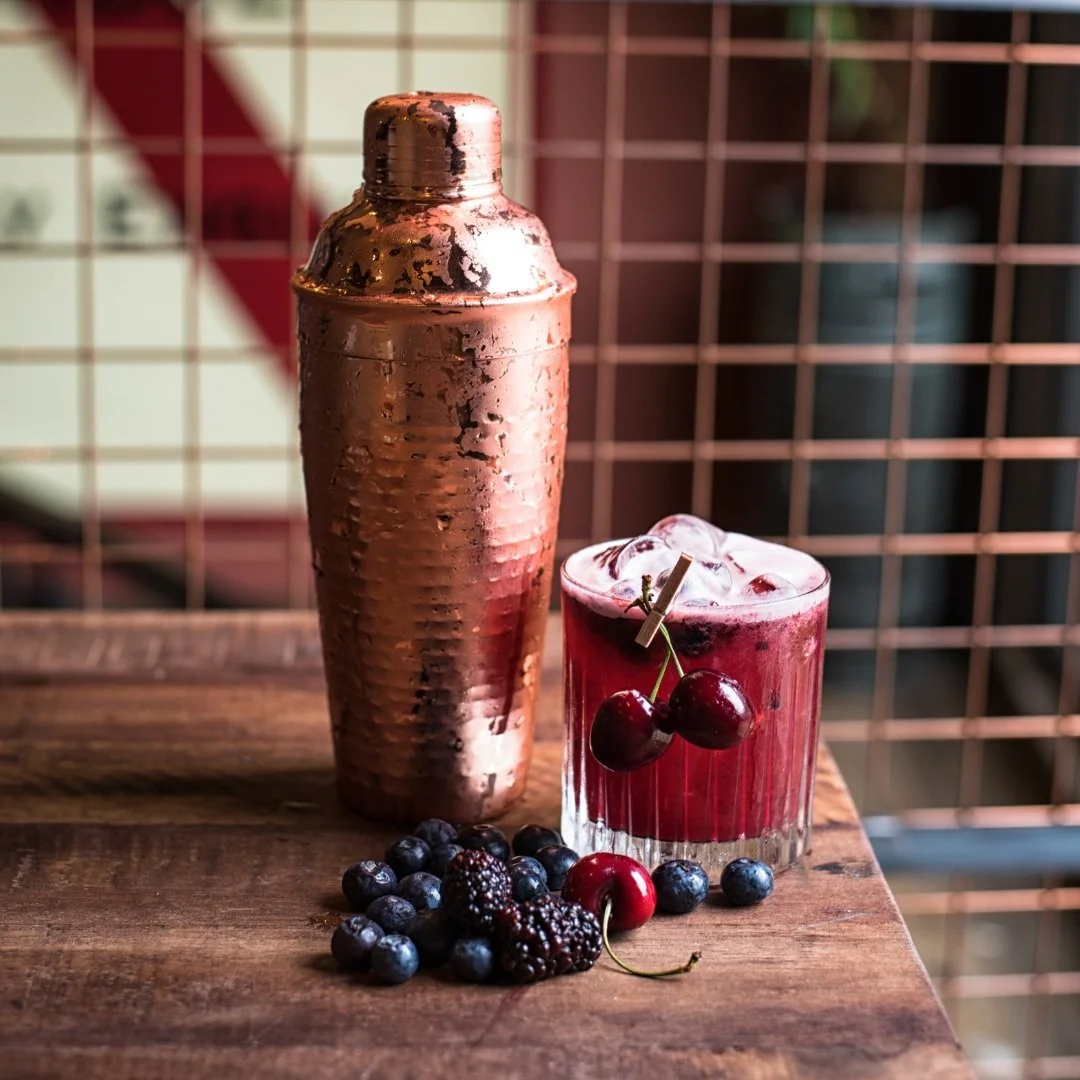 Ein Kupfer-Shaker und ein Glas mit roten Beeren und Eis, umgeben von frischen Beeren auf einem Holztisch.