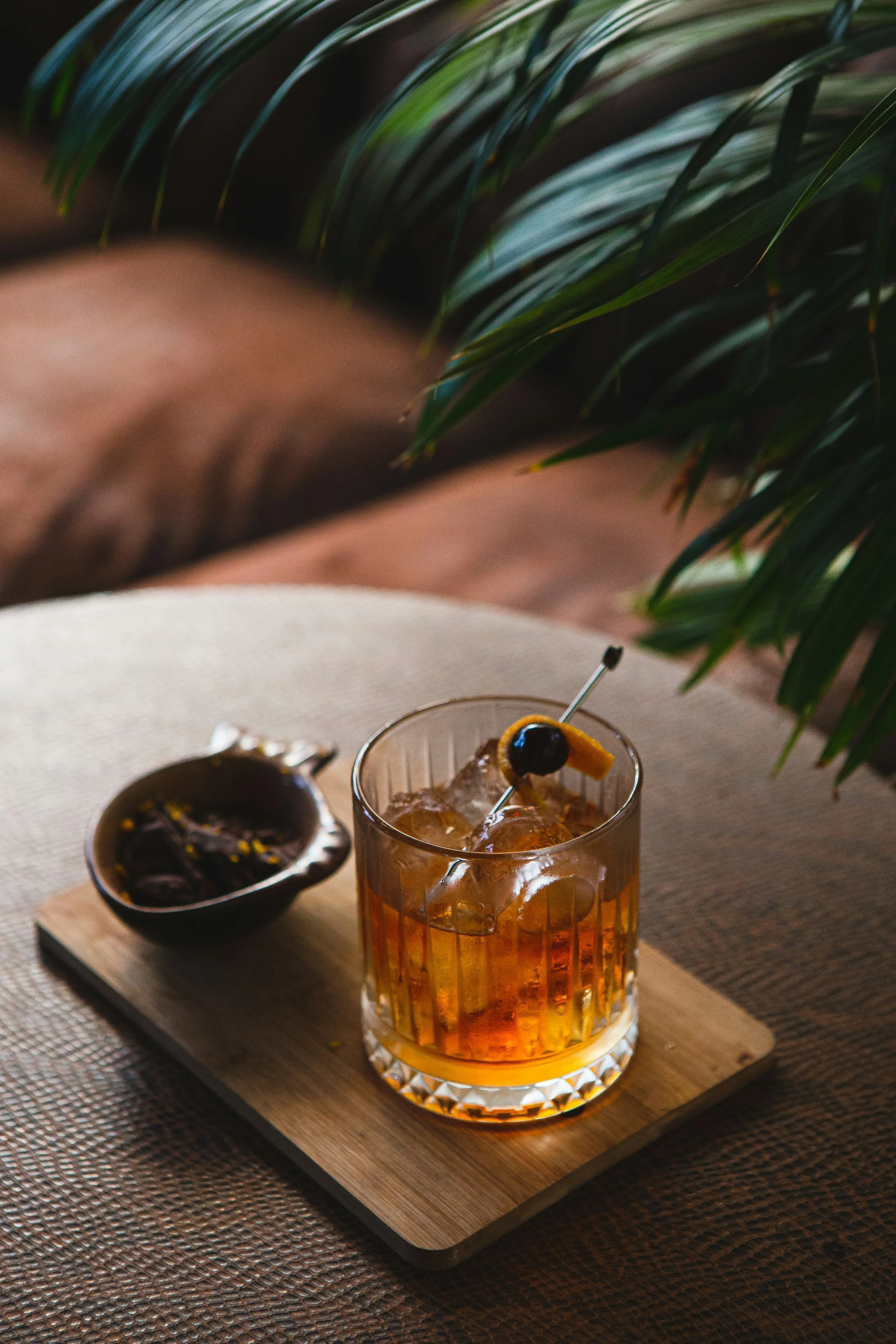 Ein Glas mit einem gelieferten Getränk, garniert mit einer Kirsche und einer Orange, auf einem Holzbrett, daneben eine kleine Schale mit dunkler Sauce und einer Gabel, auf einem Tisch mit einer gewebten Tischdecke im Hintergrund.