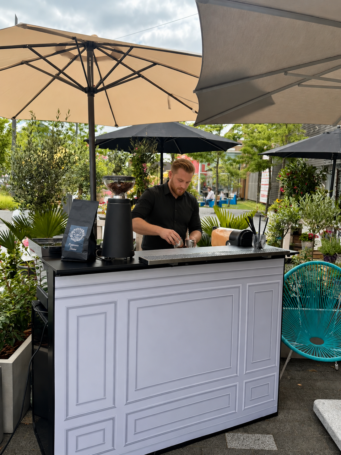 Barista bereitet an einer mobilen Kaffeebar unter Sonnenschirmen ein Getränk zu.