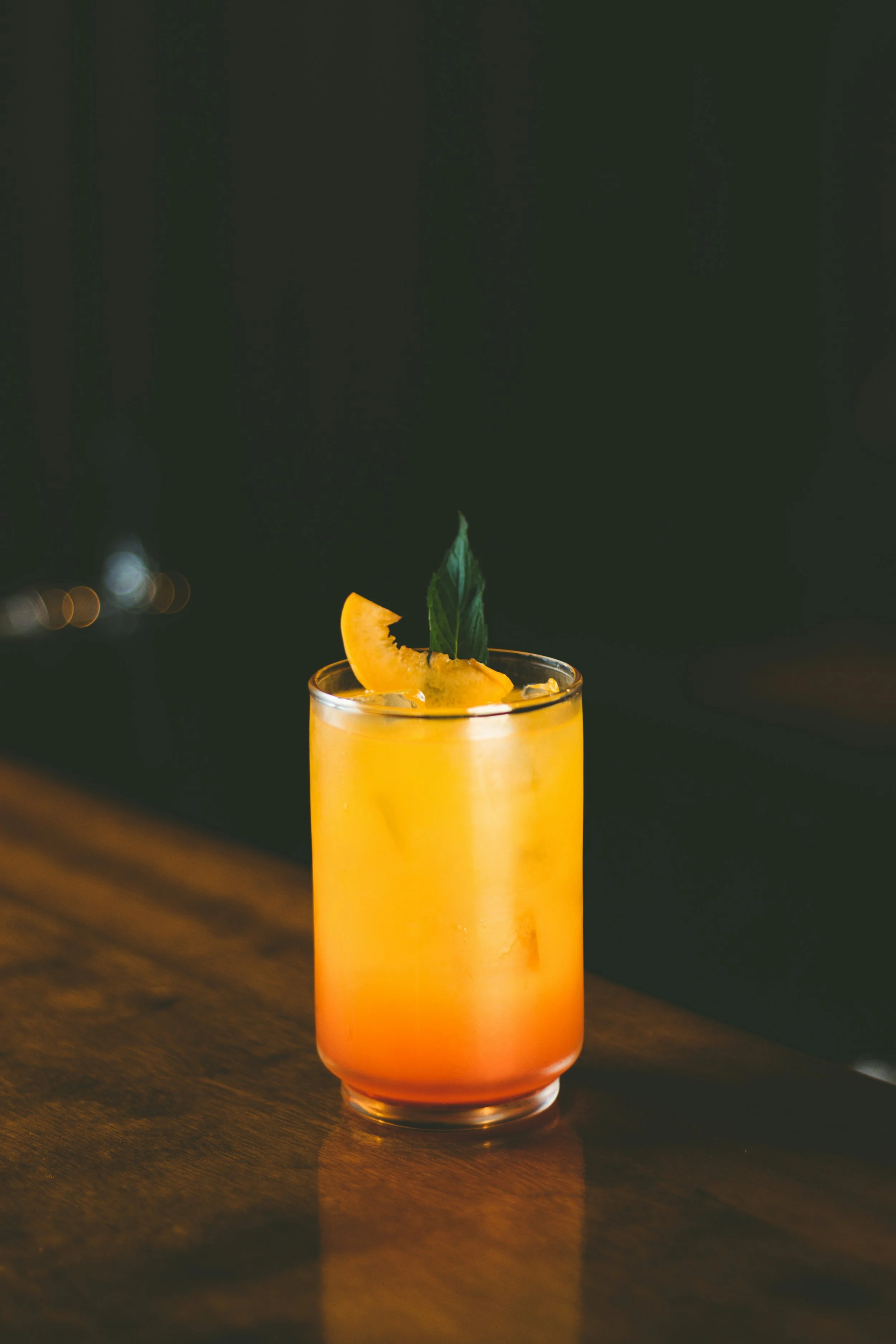 Ein hoher, bunter Cocktail in einem Glas mit einer Orangenscheibe und Minzblatt als Dekoration, auf einem dunklen Holztisch bei Nacht.
