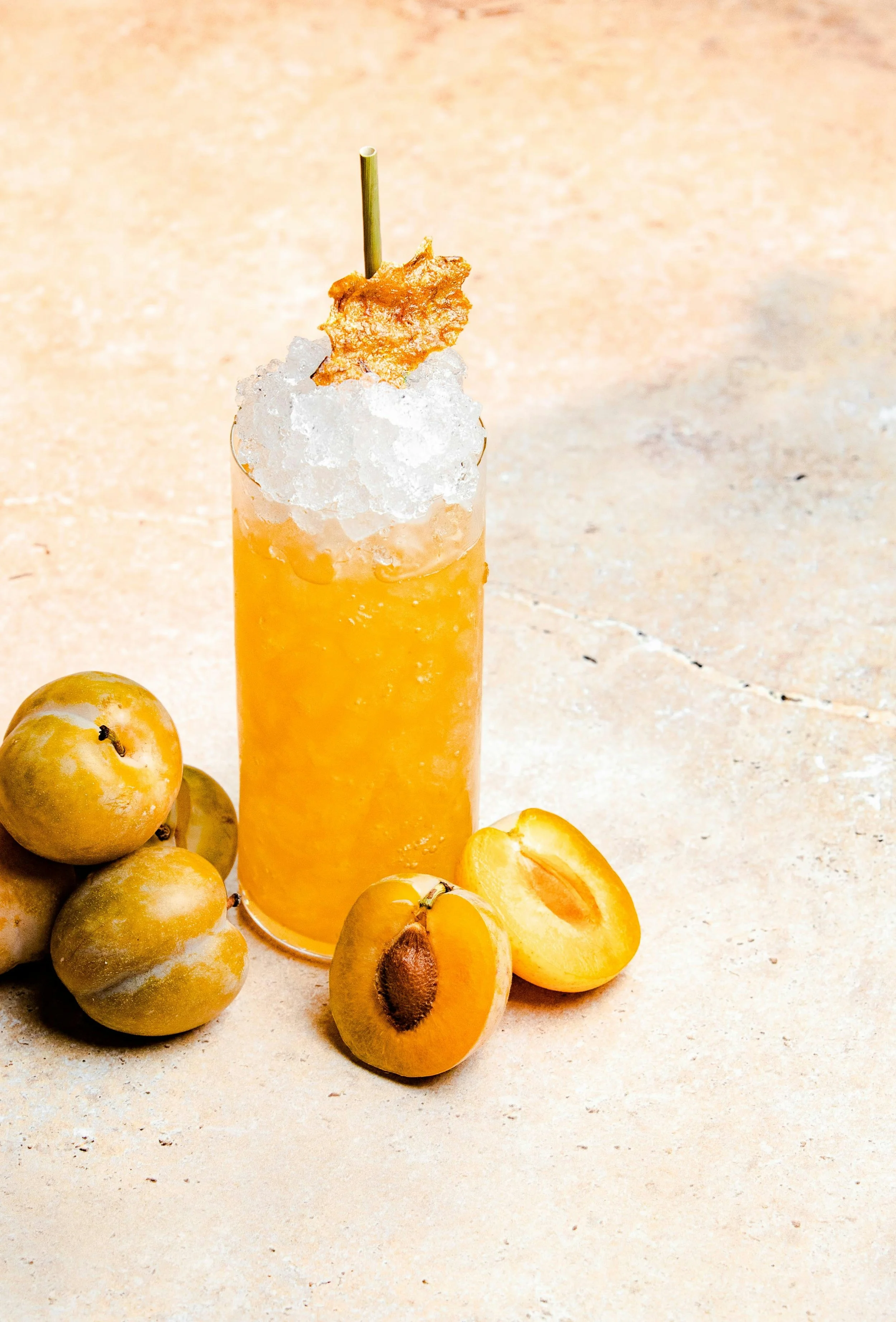 Ein sommerlicher Cocktail in einem hohen Glas, garniert mit Kokosraspeln, einem Stück gebackenen Keks und einem Strohhalm. Um das Glas herum liegen grüne, reife Aprikosen, einige halbiert, auf einer hellen Oberfläche.