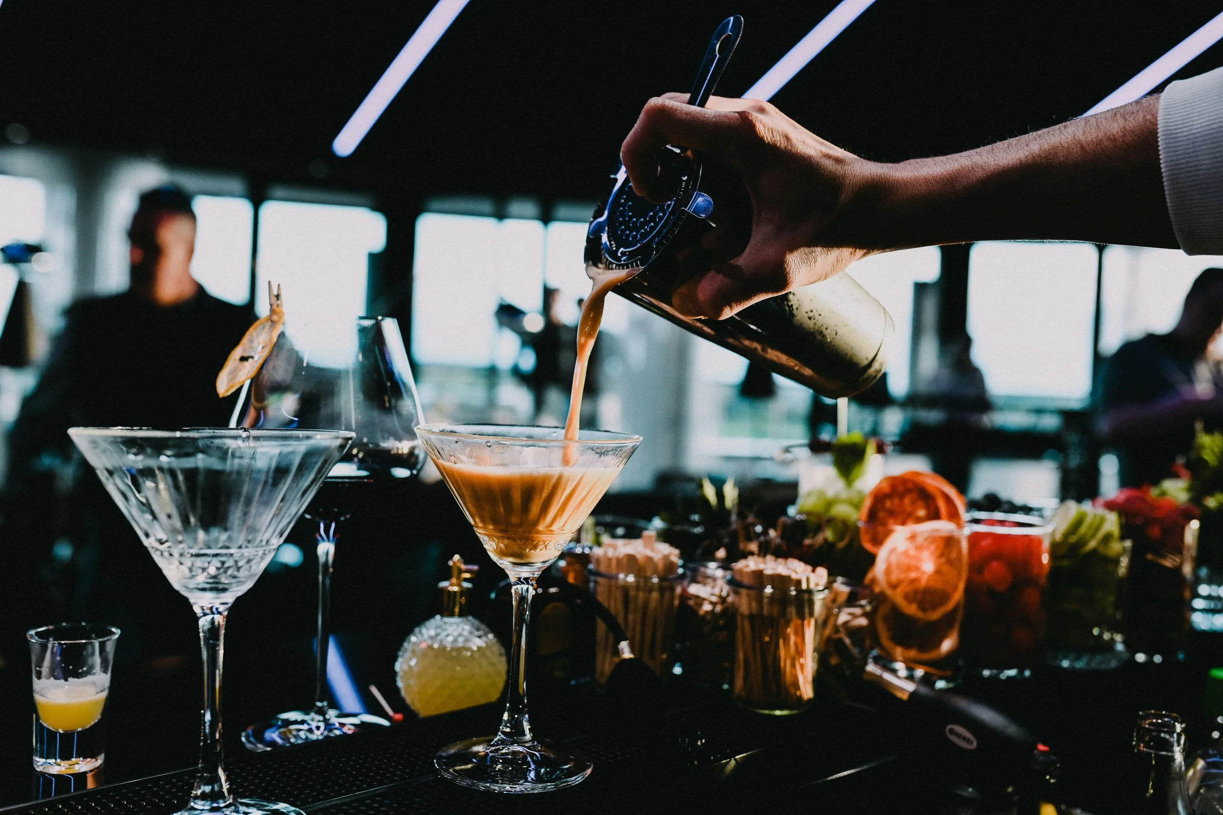 Hand gießt braune Flüssigkeit in ein Cocktailglas bei einer Veranstaltung mit dunklem Hintergrund und Menschen im Hintergrund.