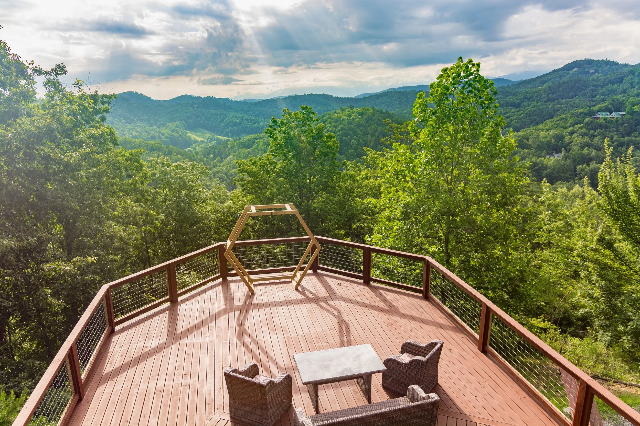 A wooden deck with a table and chairs overlooking a lush green mountain landscape under a cloudy sky.