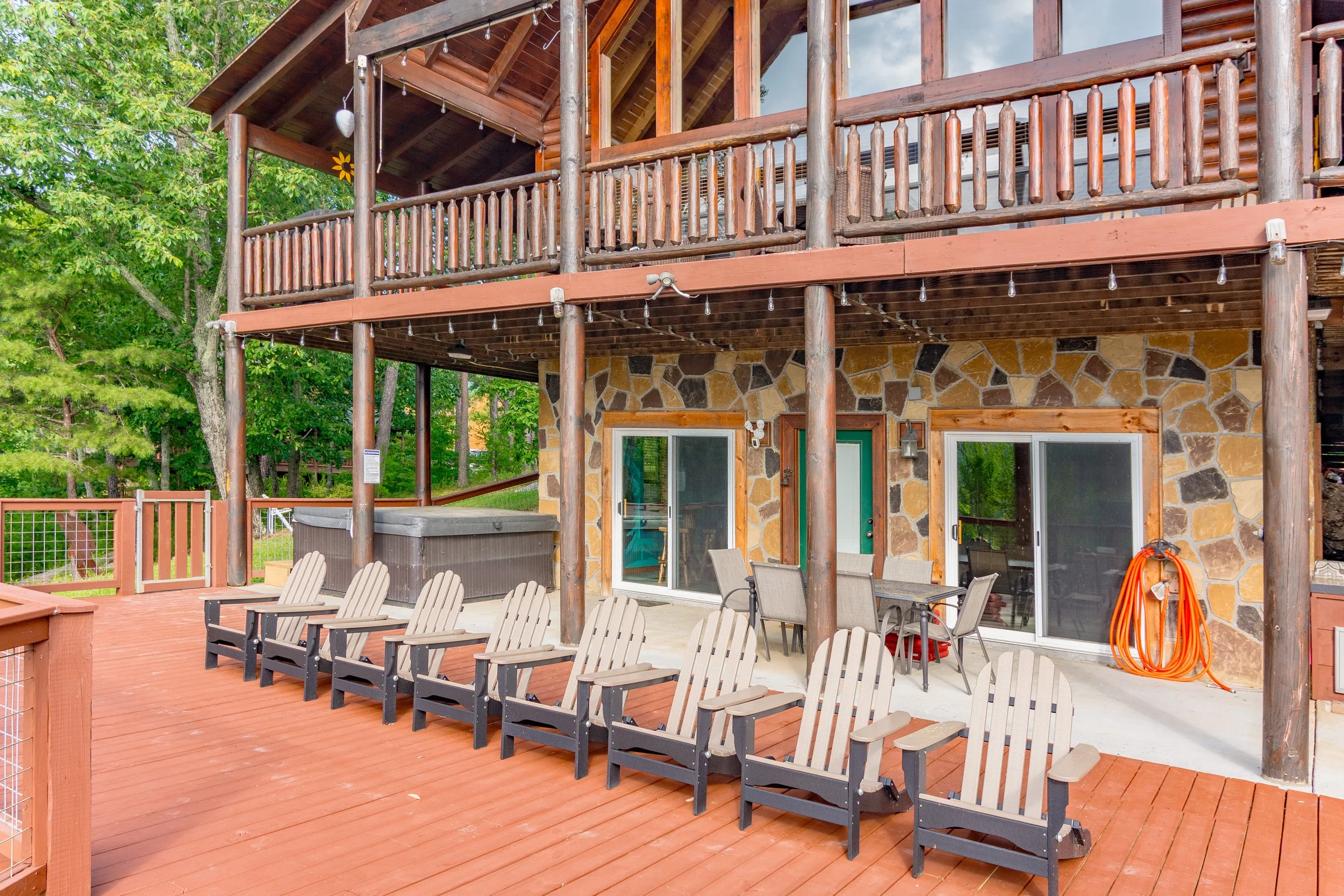 A wooden deck attached to a house with stone walls, featuring Adirondack chairs, a hot tub, a dining table with chairs, and an orange garden hose. The house has a balcony with wooden railings, and there are trees in the background.