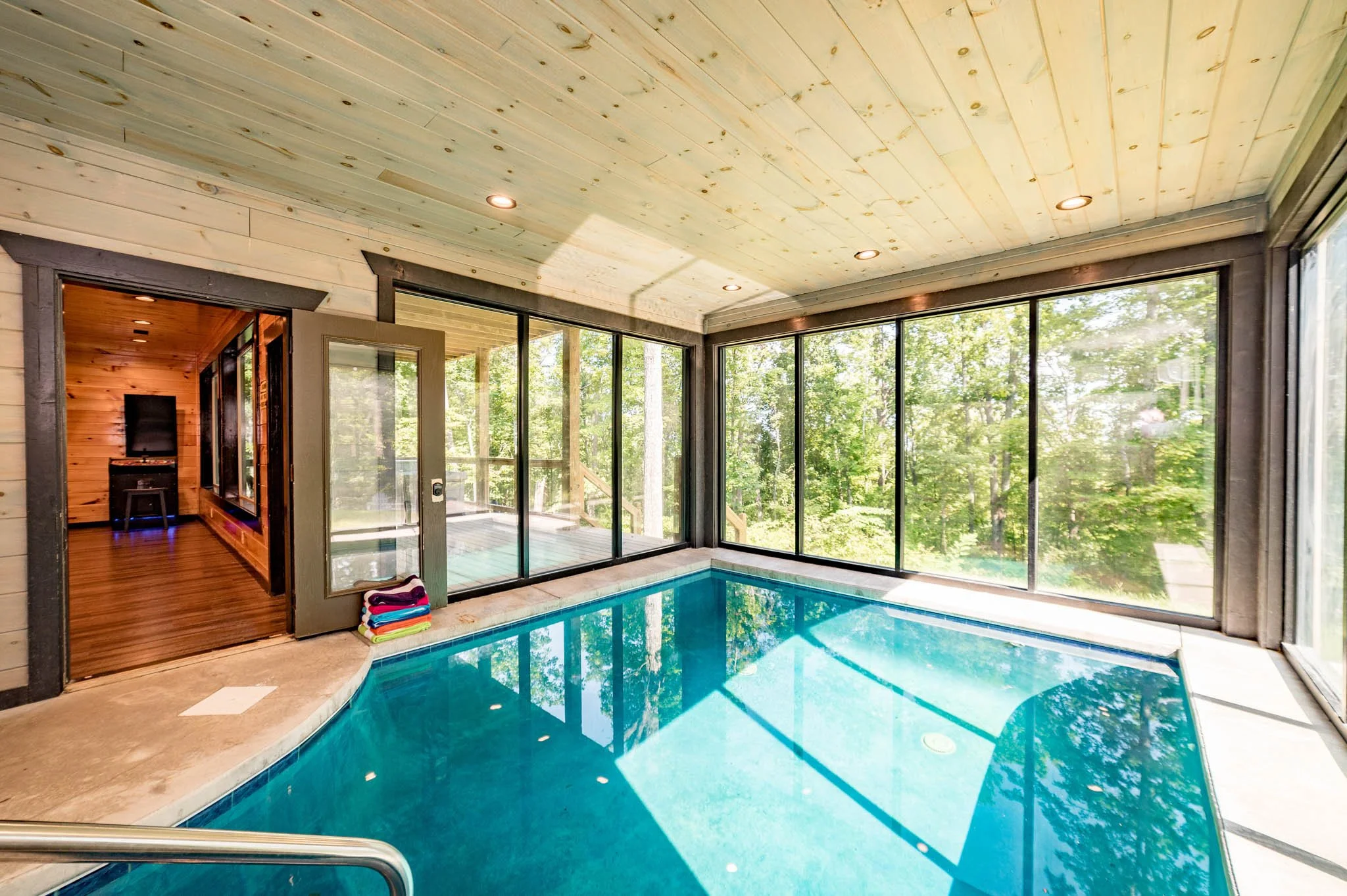 Indoor swimming pool with large glass windows showing lush green trees outside, wood-paneled ceiling with built-in lights, and colorful towels stacked on the ledge near the pool.