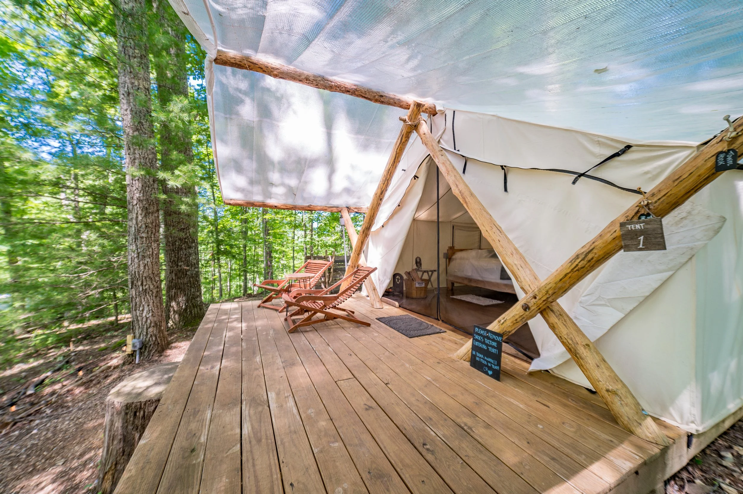 Wooden tent platform with two Adirondack chairs outside a canvas tent in a forested area with green trees.