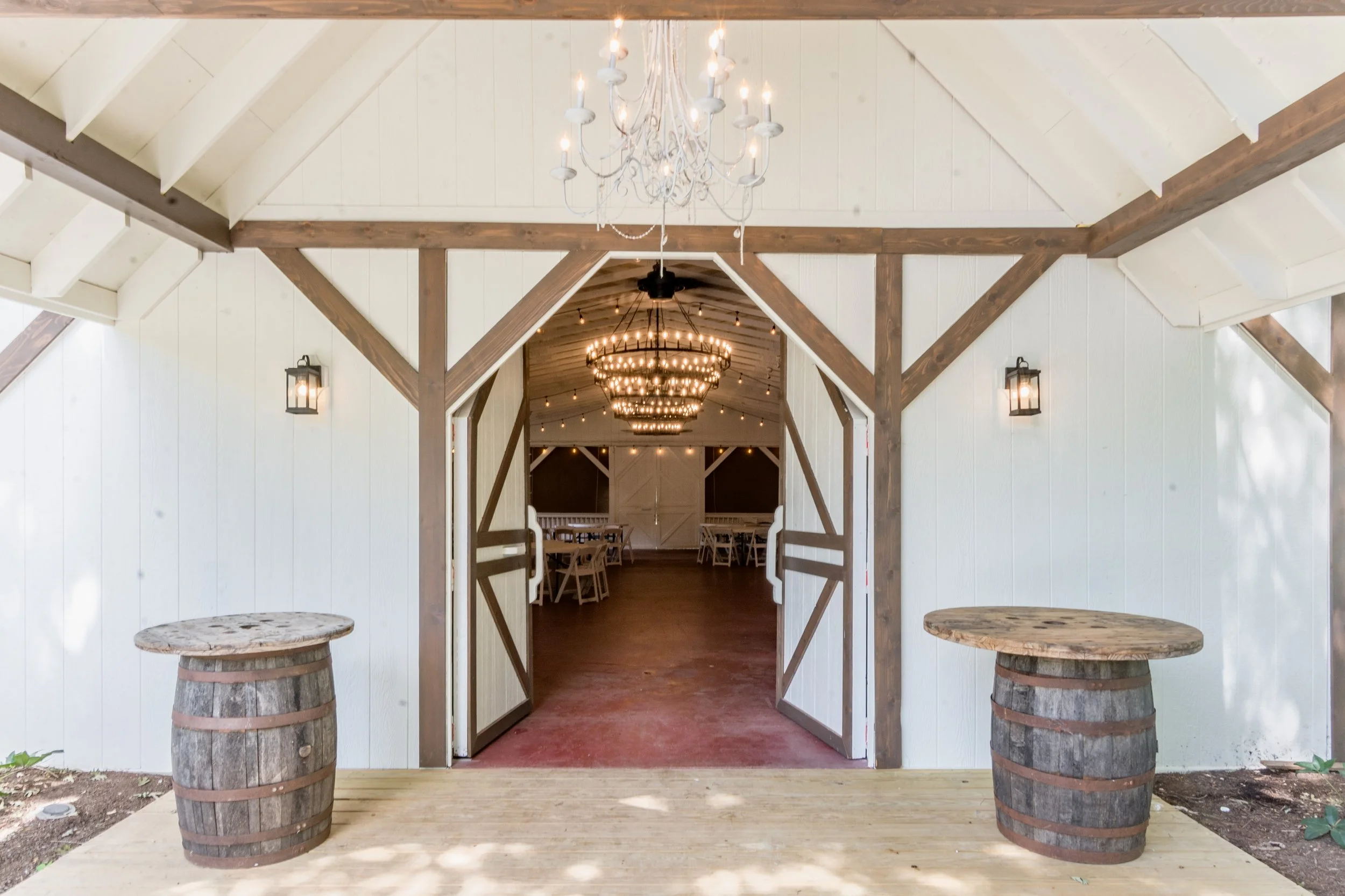 Interior of a rustic barn event space with white walls, exposed wood beams, a chandelier, and round tables with chairs inside.