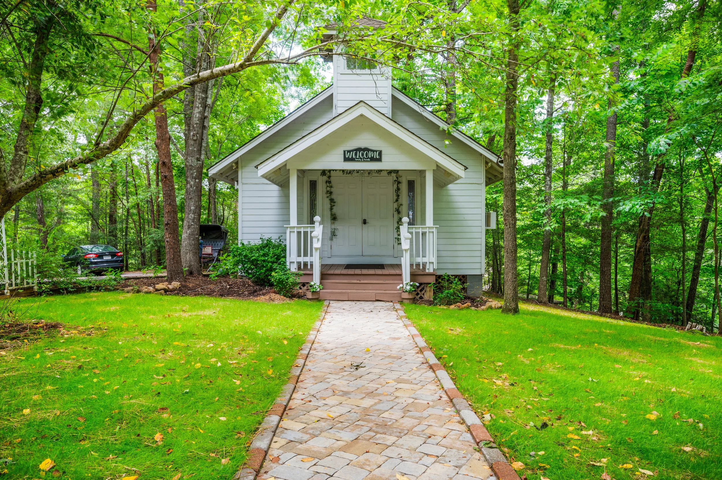 White house with a front porch, small stairs, and a sign that says "Welcome" on the porch, surrounded by green trees and grass.
