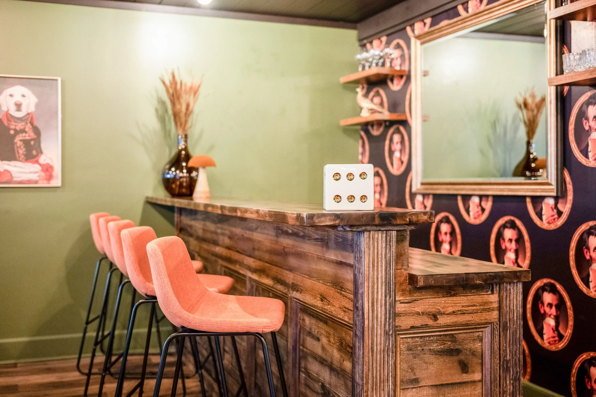 Interior of a bar with a green wall, a wooden counter, pink bar stools, a framed dog photo, decorative vases, and a large mirror.