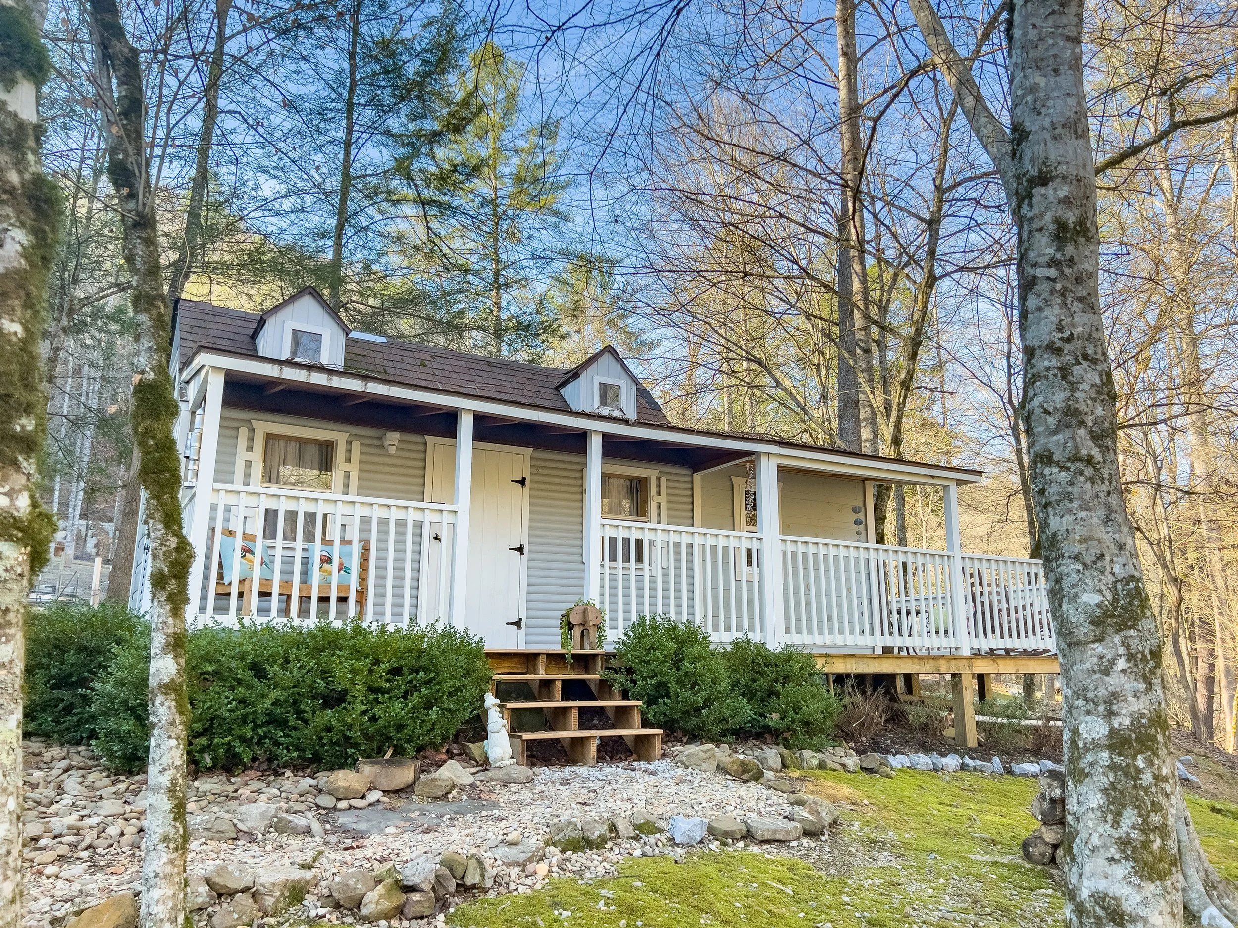 Small white house with a covered porch, surrounded by trees and bushes, in a wooded area.