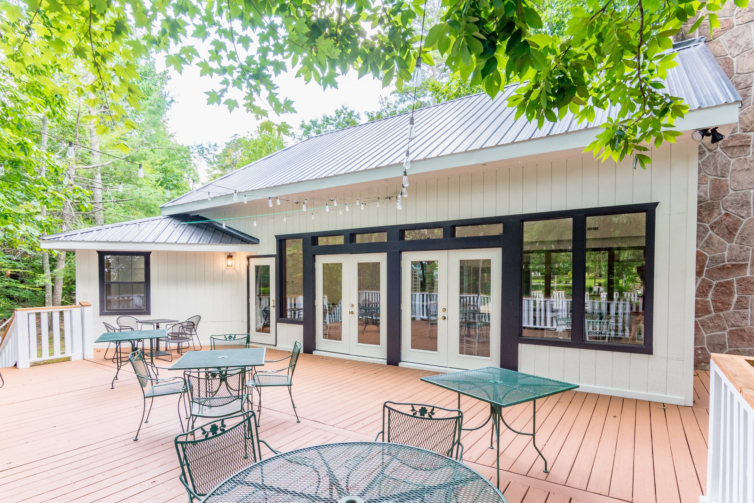 Exterior view of a backyard patio with metal tables and chairs, a white house with black trim, large windows, a door, string lights hanging above, surrounded by green trees.