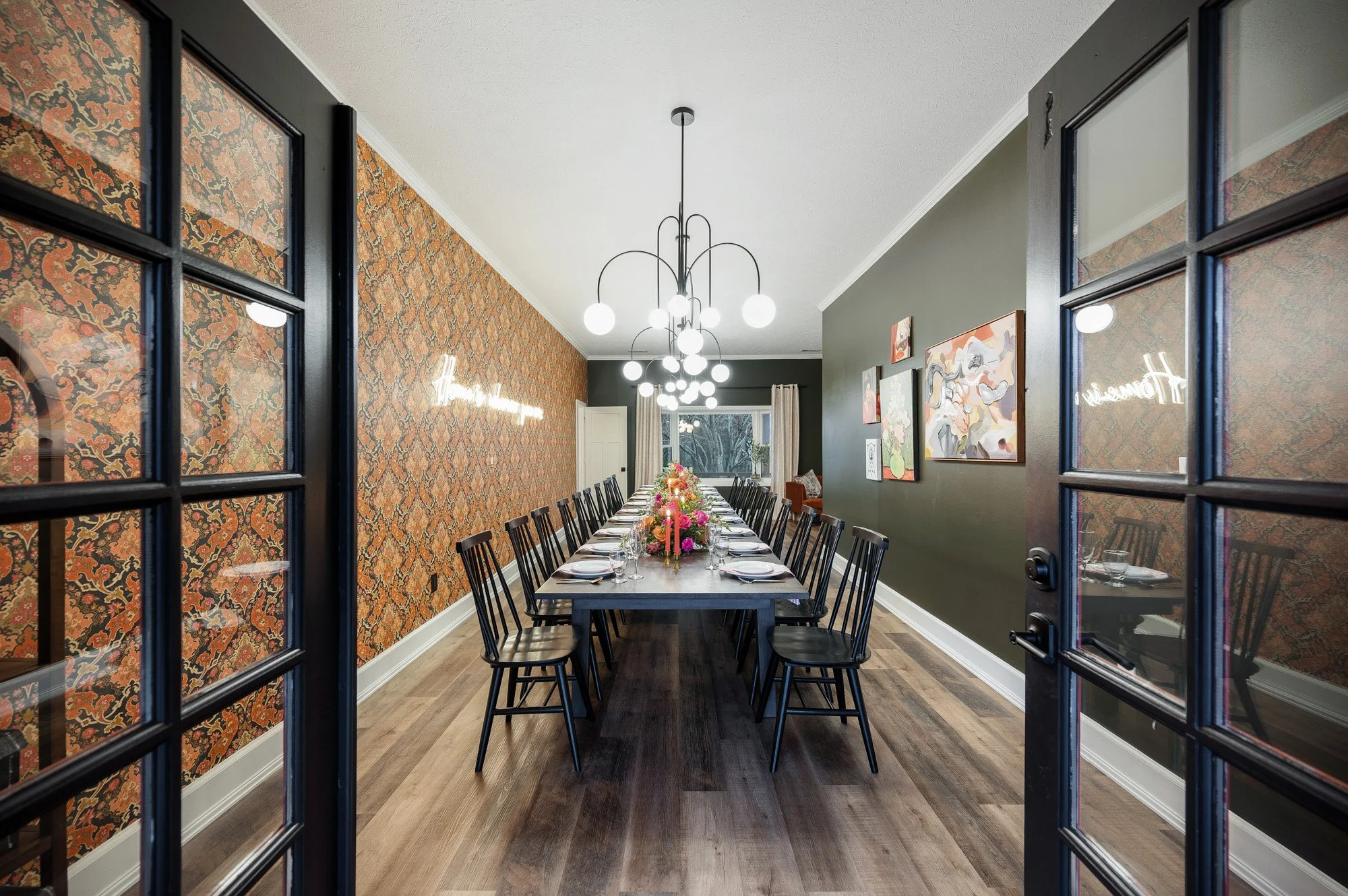 A dining room with a long table set for a meal, featuring a floral centerpiece, surrounded by black chairs, with patterned wallpaper on one wall and artwork on another. A modern chandelier hangs above the table, and french doors are on the sides.