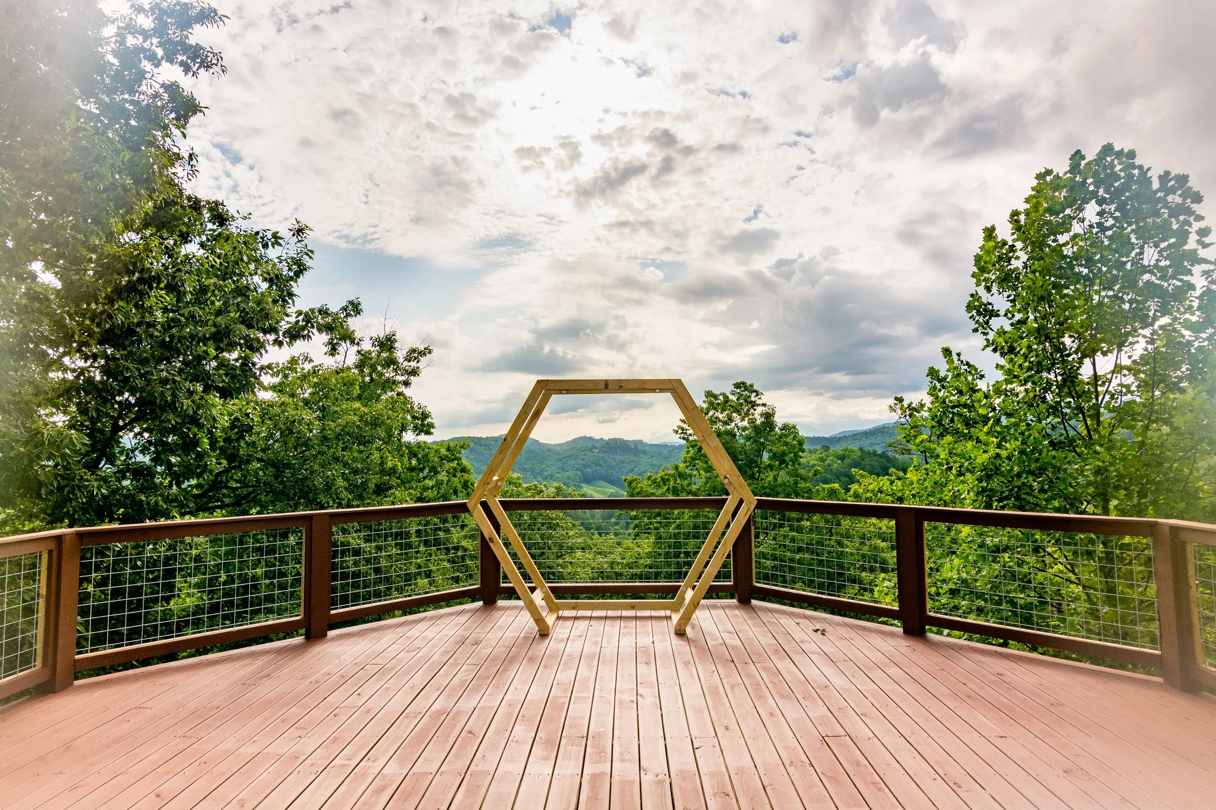 A wooden deck with a hexagonal wooden frame against a scenic backdrop of lush green trees and distant hills under a partly cloudy sky.