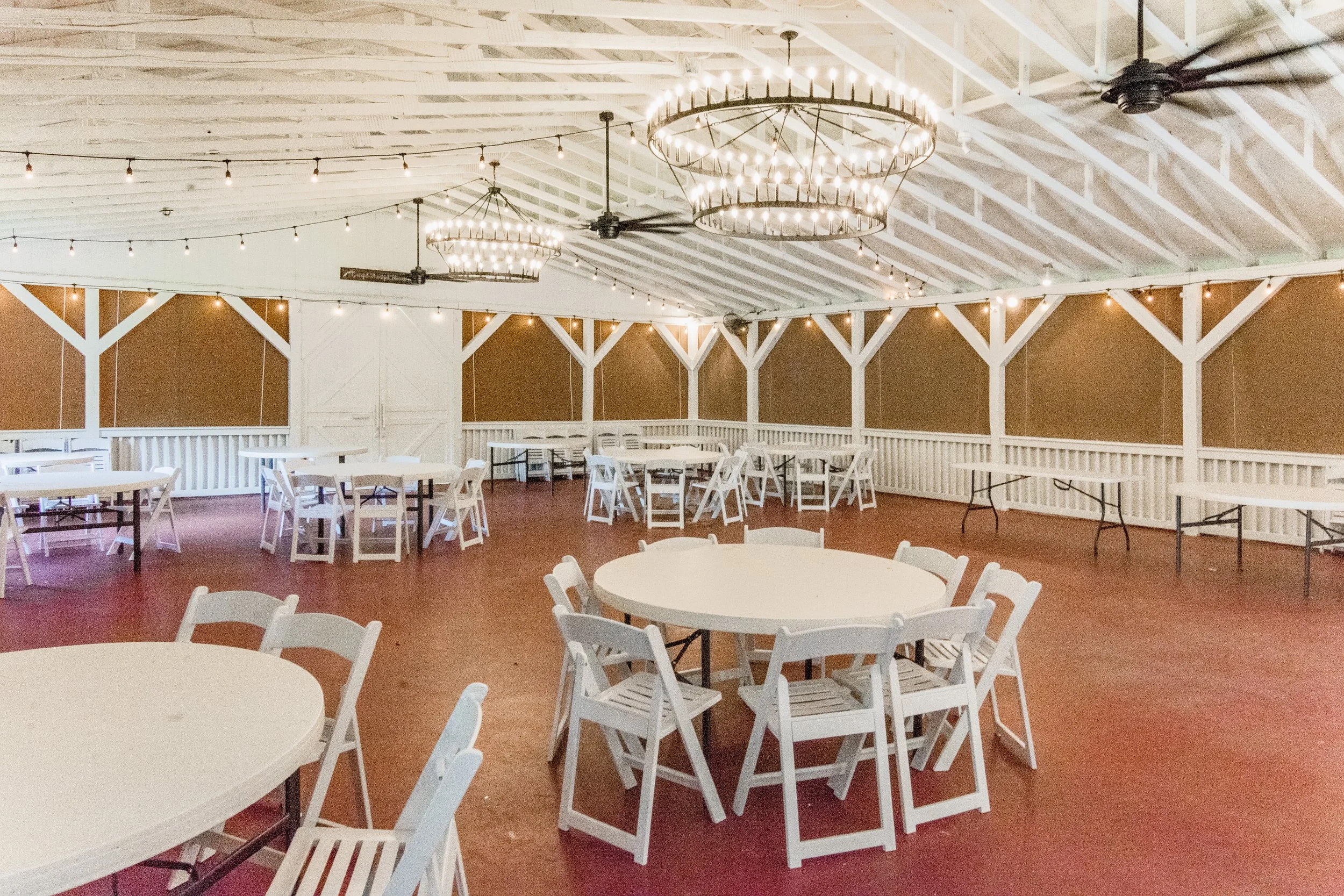 Empty event space with round tables and chairs, string lights, ceiling fans, and large chandeliers in a rustic hall.