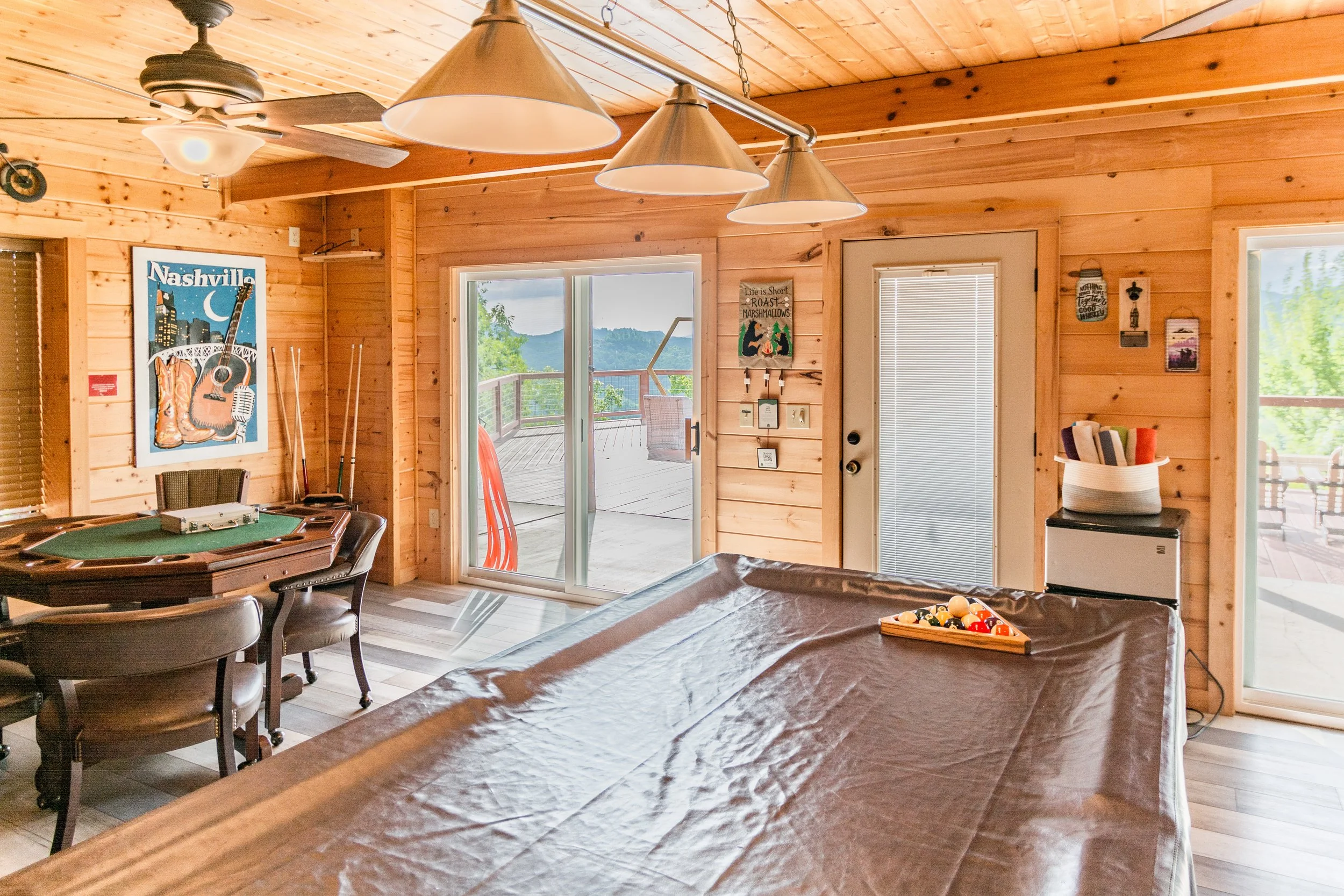 Interior view of a wooden cabin game room with a pool table covered with a brown cloth, a dartboard, a corner with a small fridge, and a sliding glass door leading to a deck with mountain views.